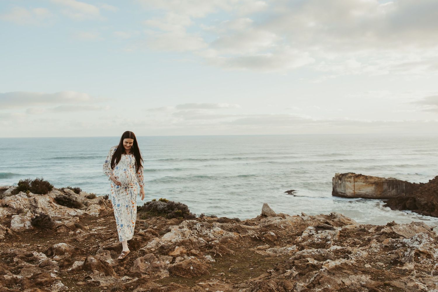 Woman in white lace outfit walking on rocky coastal cliffs with ocean and cloudy sky in background.