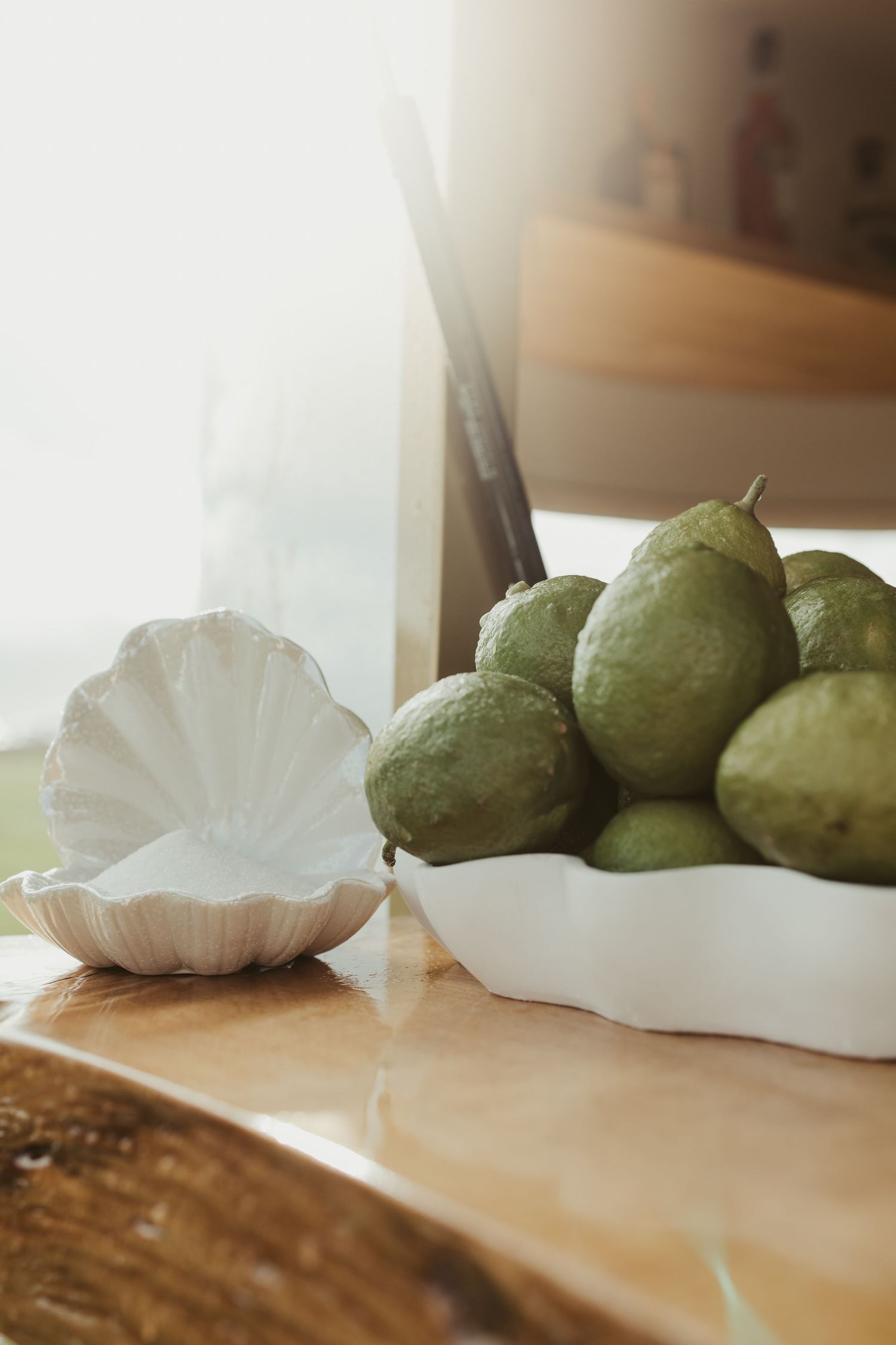 Fresh green guavas in white scalloped ceramic dish beside white shell bowl on wooden table in sunlight.