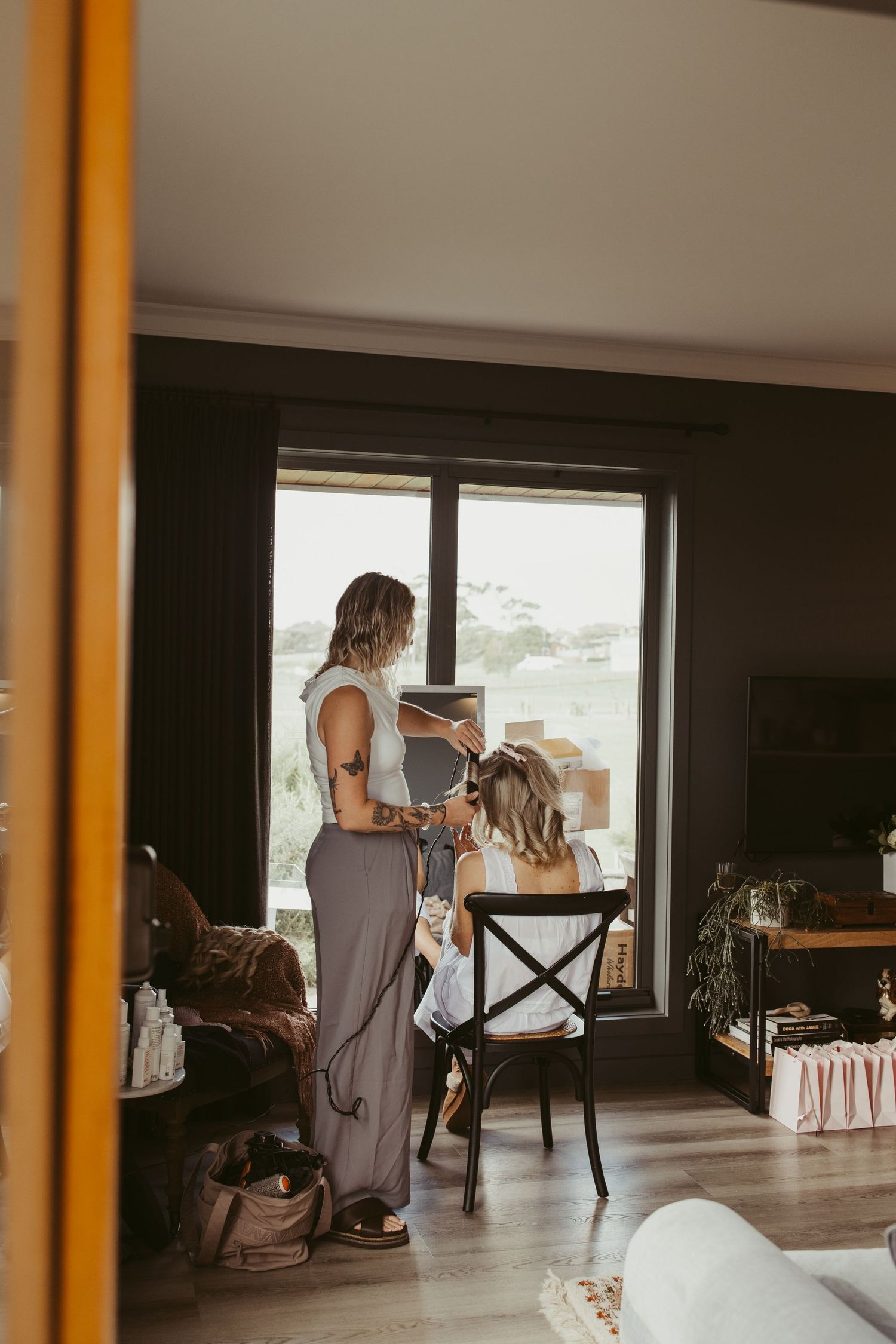 Hair stylist styling seated bride's hair near large window in modern dark-toned bridal suite.