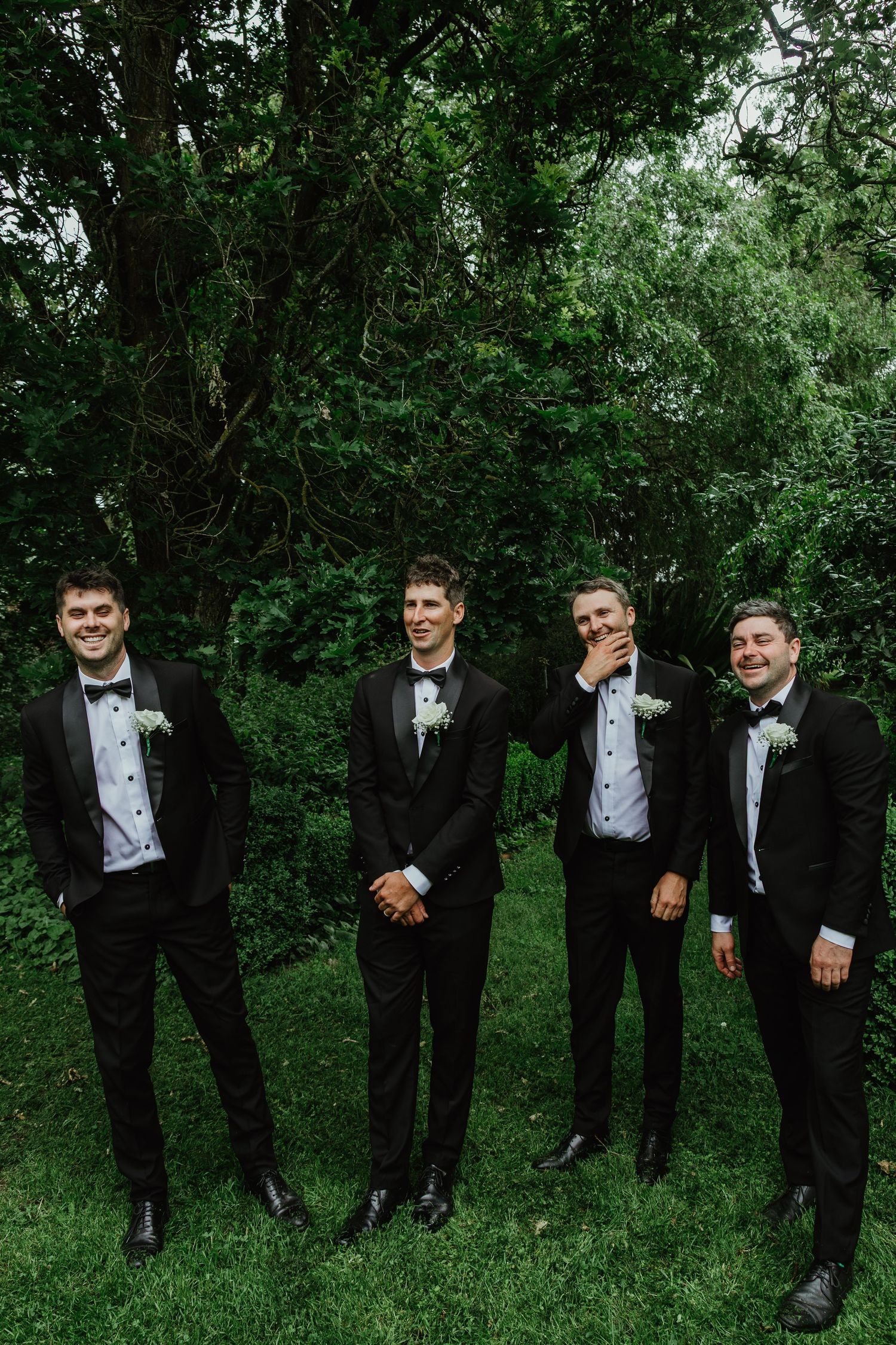 Four groomsmen in black tuxedos with bow ties laughing together in front of lush green trees.