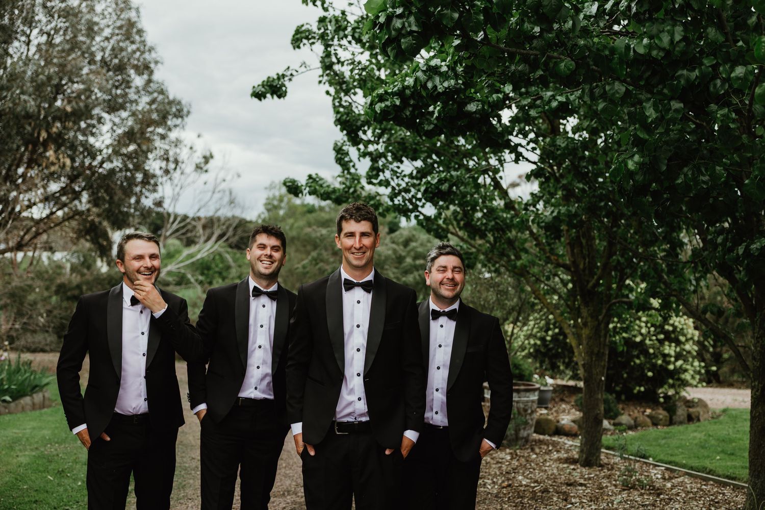 Four groomsmen in black tuxedos with bow ties smiling outdoors in a garden setting on a wedding day.