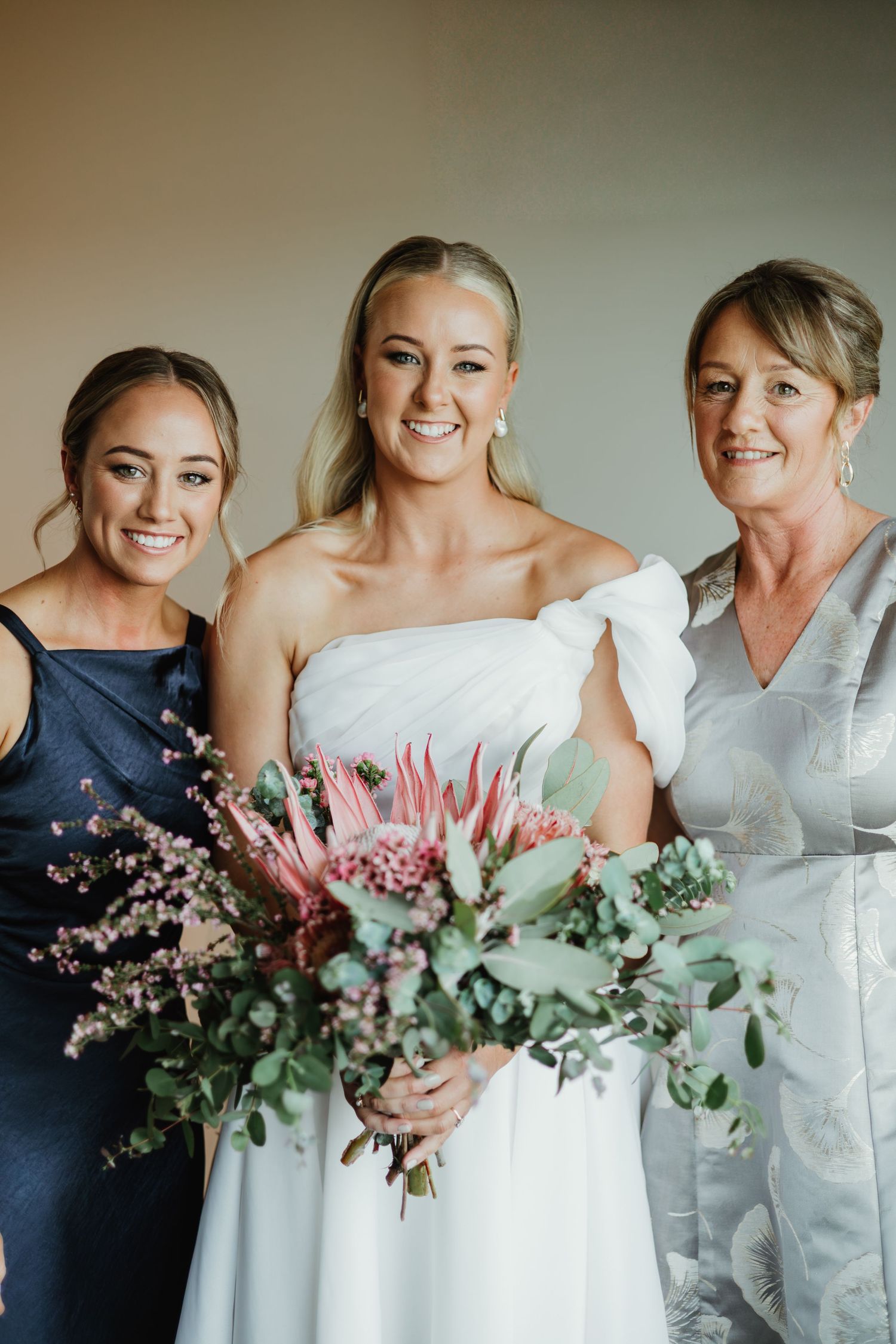 Bride in white strapless gown holds protea bouquet flanked by bridesmaid and mother.