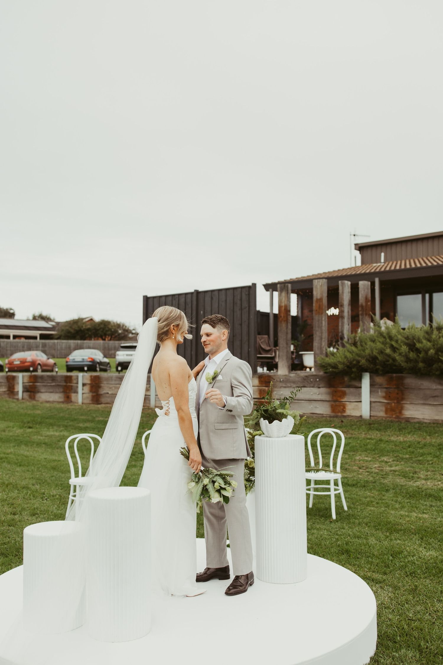 Bride and groom standing together on white circular ceremony platform surrounded by white chairs and florals.
