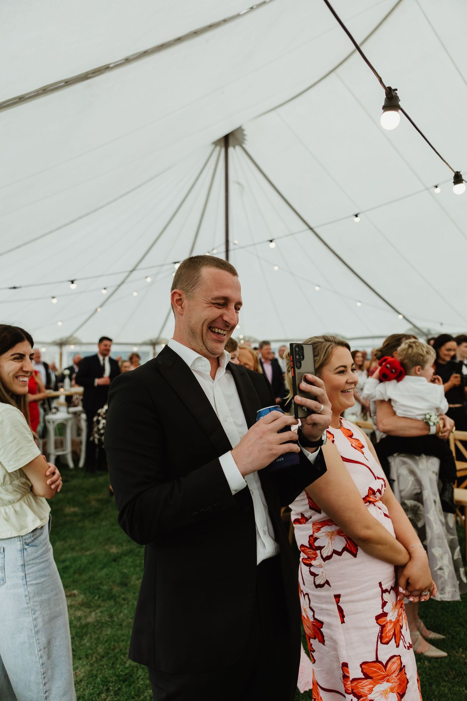 Wedding guests laughing and holding drinks under white tent with string lights at reception.