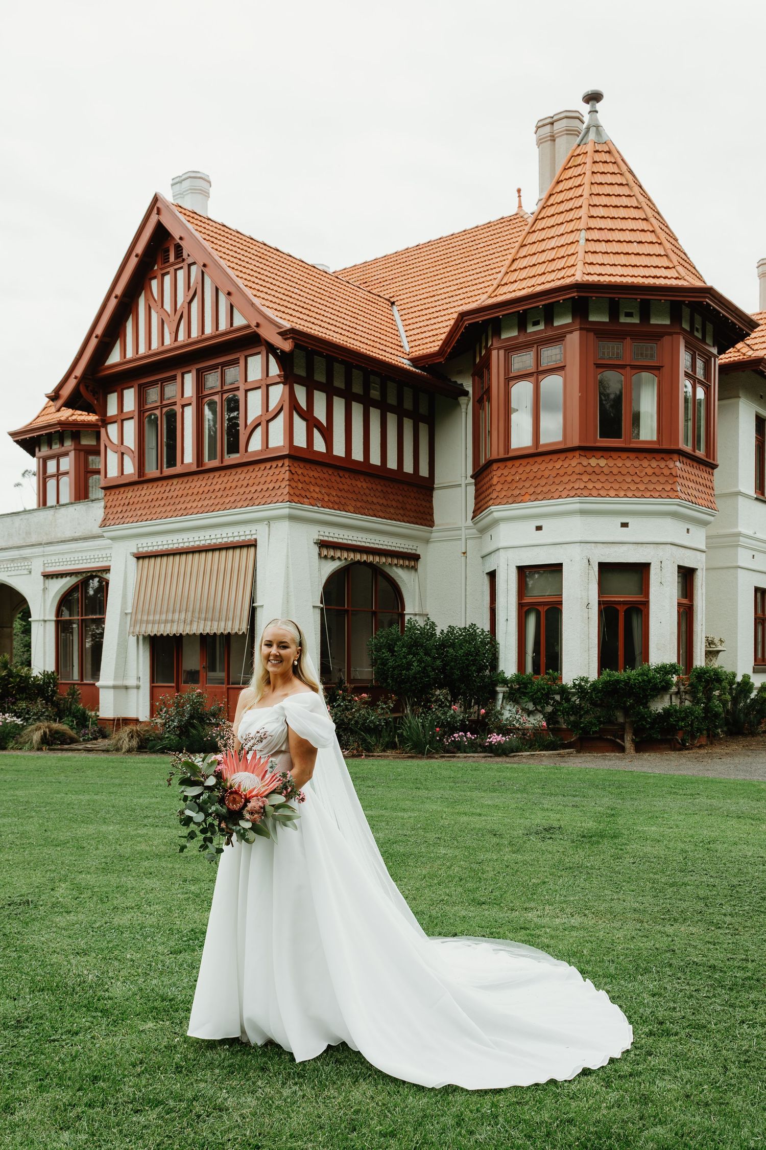 Bride in white off-shoulder gown holding a protea bouquet before a Tudor-style mansion.