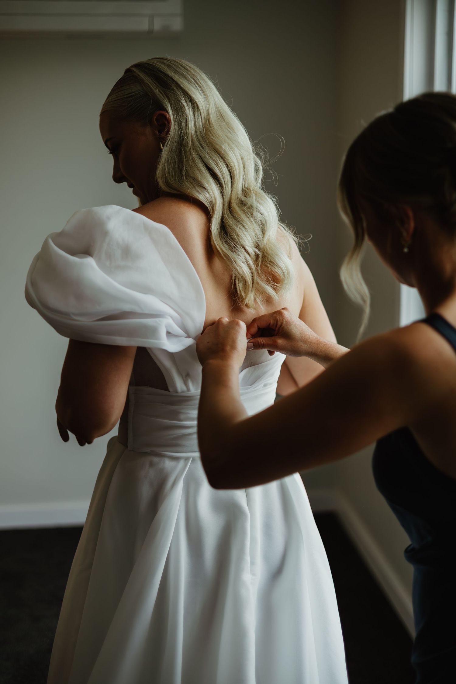 Bridesmaid fastens buttons on the back of a brides white off-shoulder puff sleeve wedding gown.