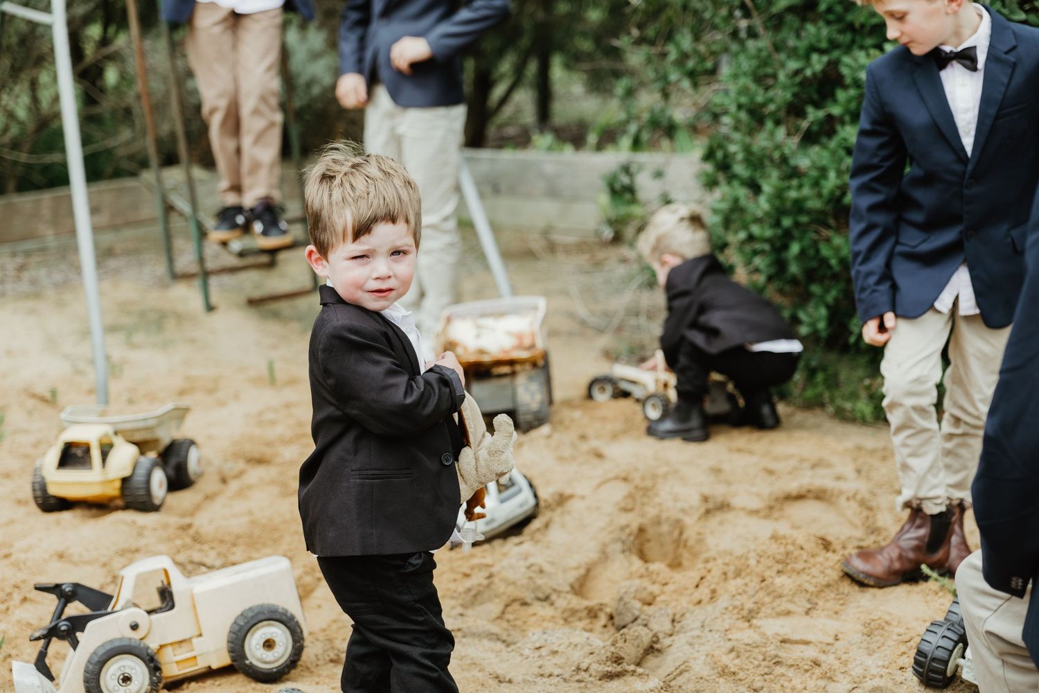 Young boy in black suit playing in sandbox with toy trucks at outdoor children's party.