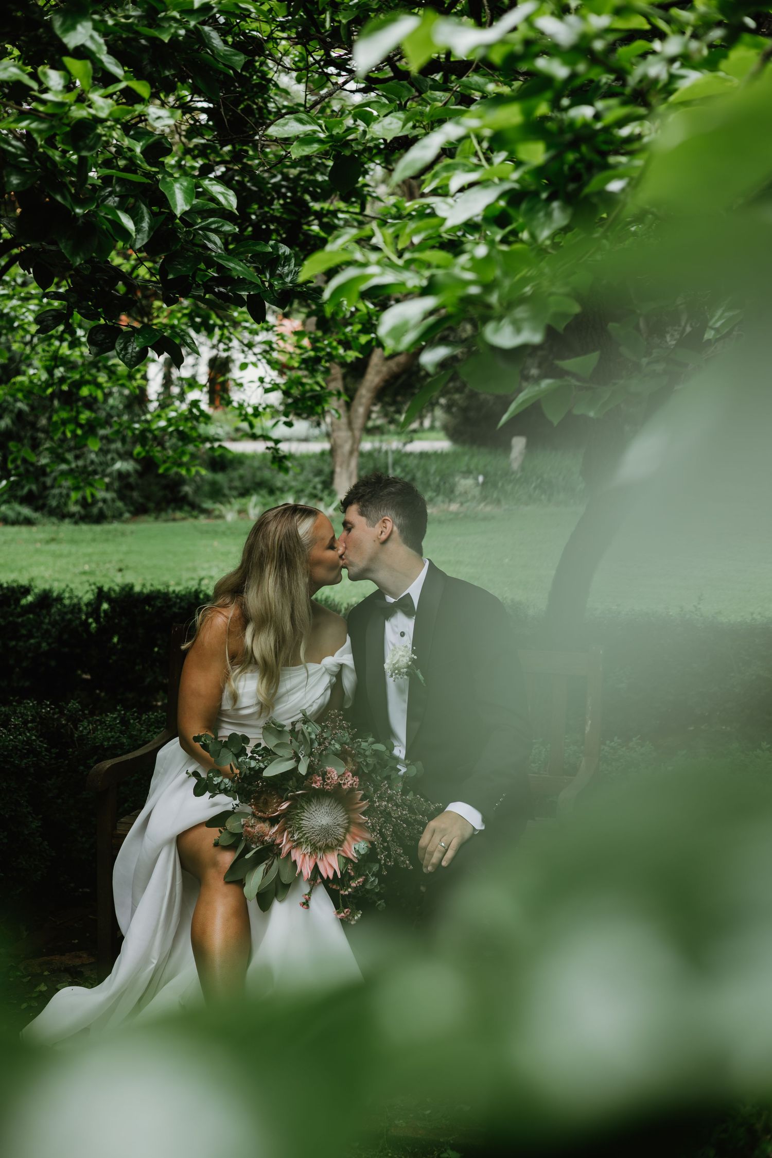Bride and groom sharing a romantic kiss beneath lush green trees in a garden wedding setting.