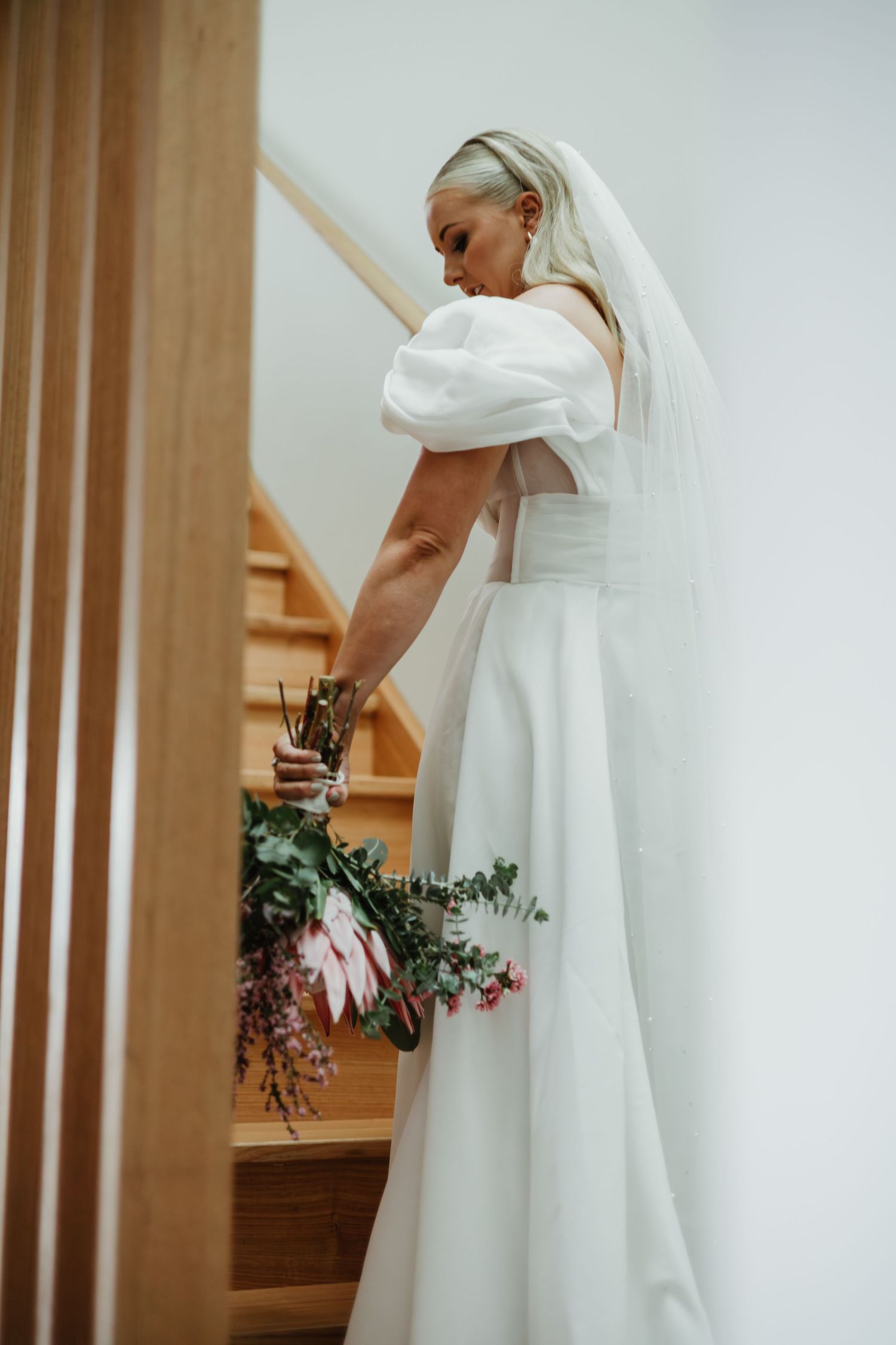 Bride in white off-shoulder gown with veil holds bouquet on wooden staircase.