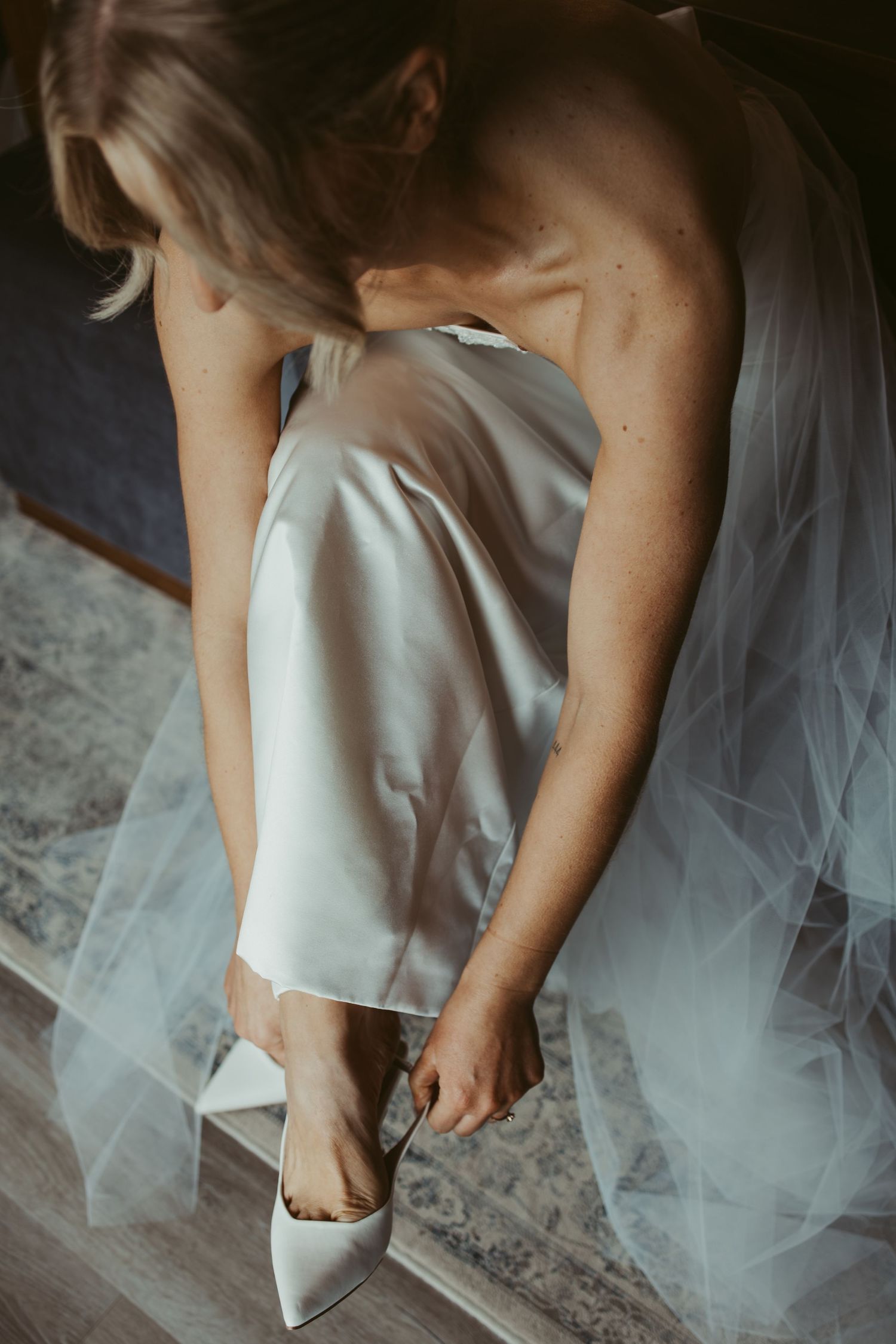 Bride in white satin slip dress adjusting her pointed-toe heels, with tulle veil draped around her.