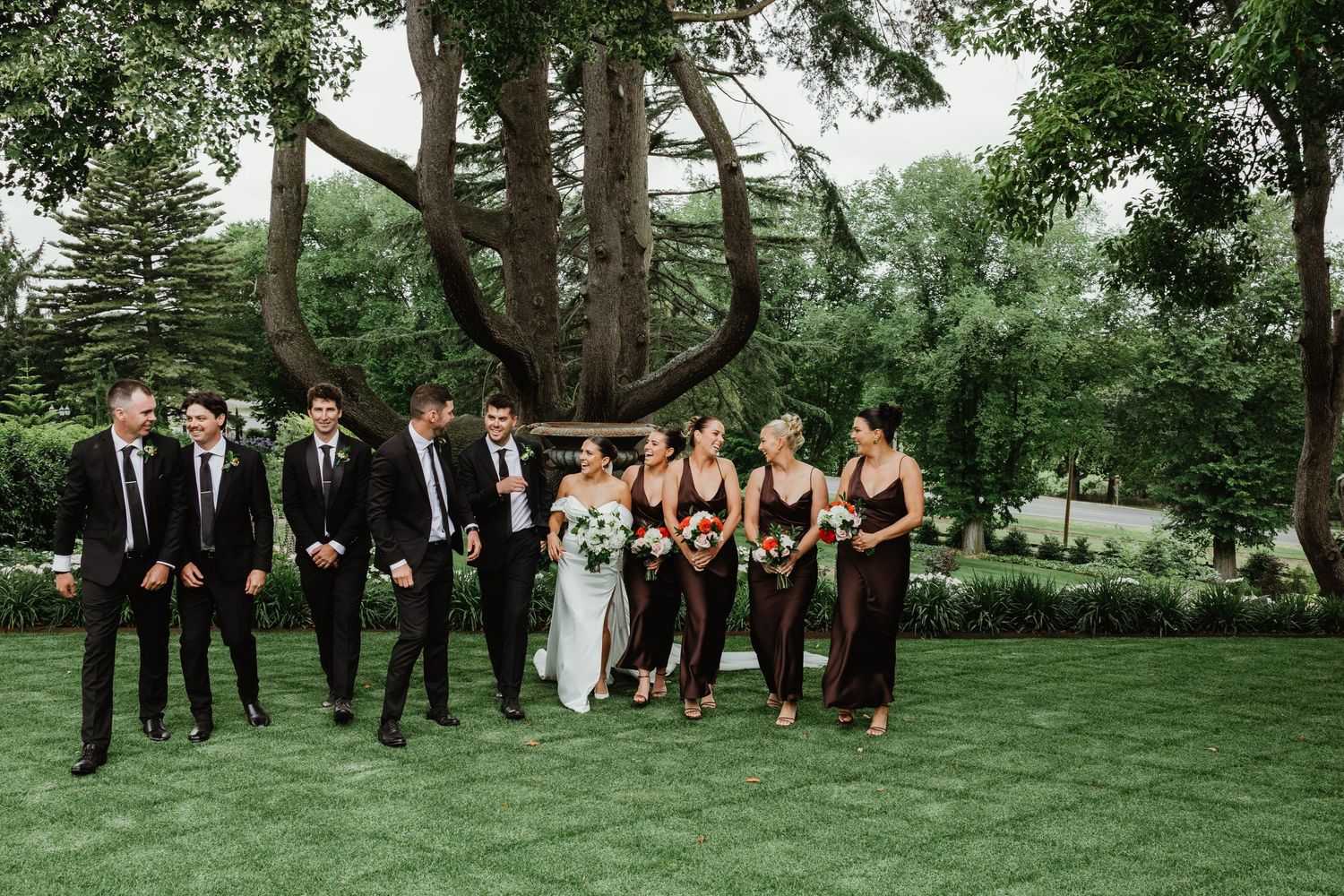 Wedding party walks on lush green lawn, groomsmen in black suits and bridesmaids in dark gowns, with lush trees behind.