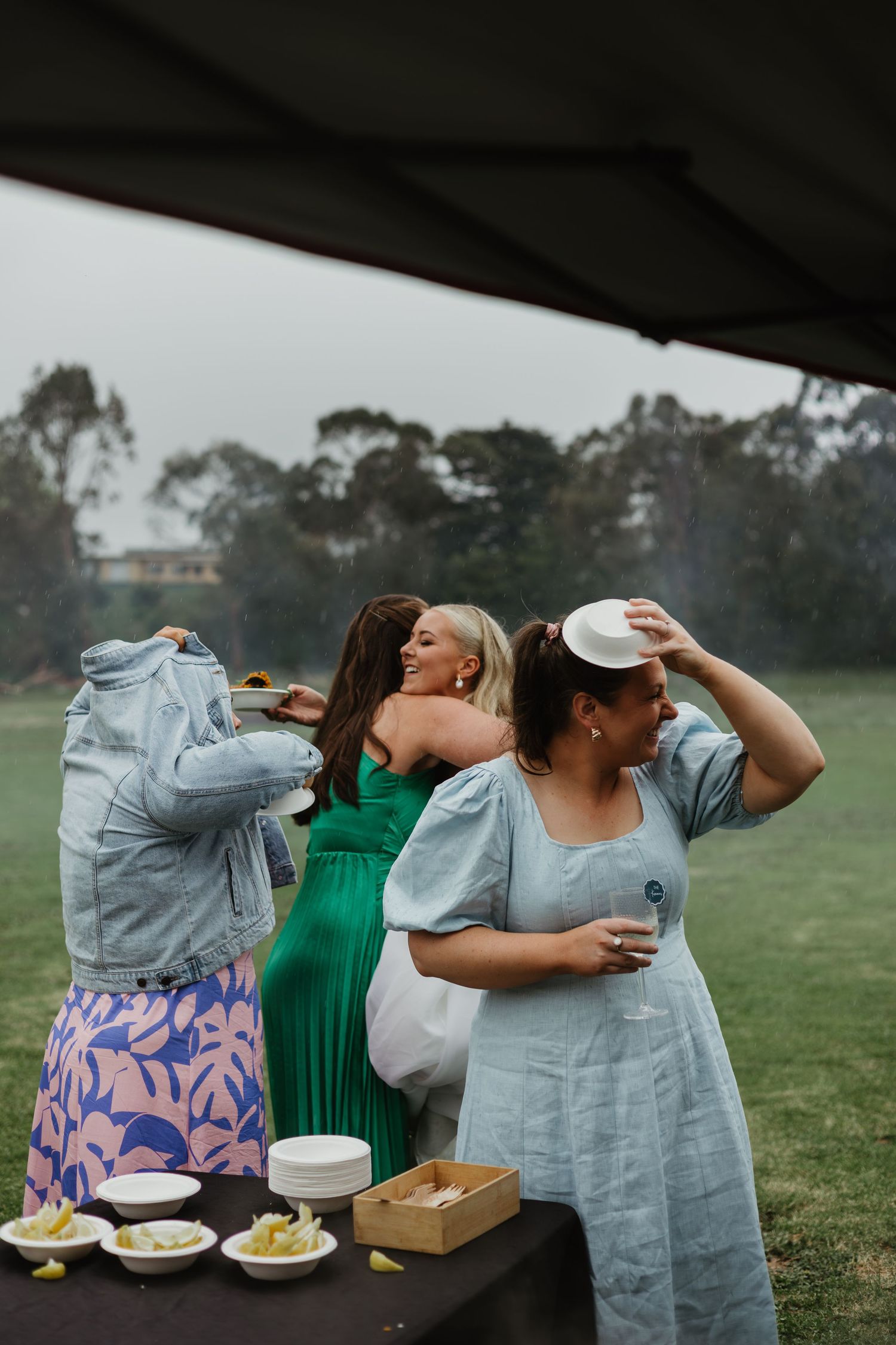 Three women in colorful dresses laughing and holding hats at a breezy outdoor wedding reception.