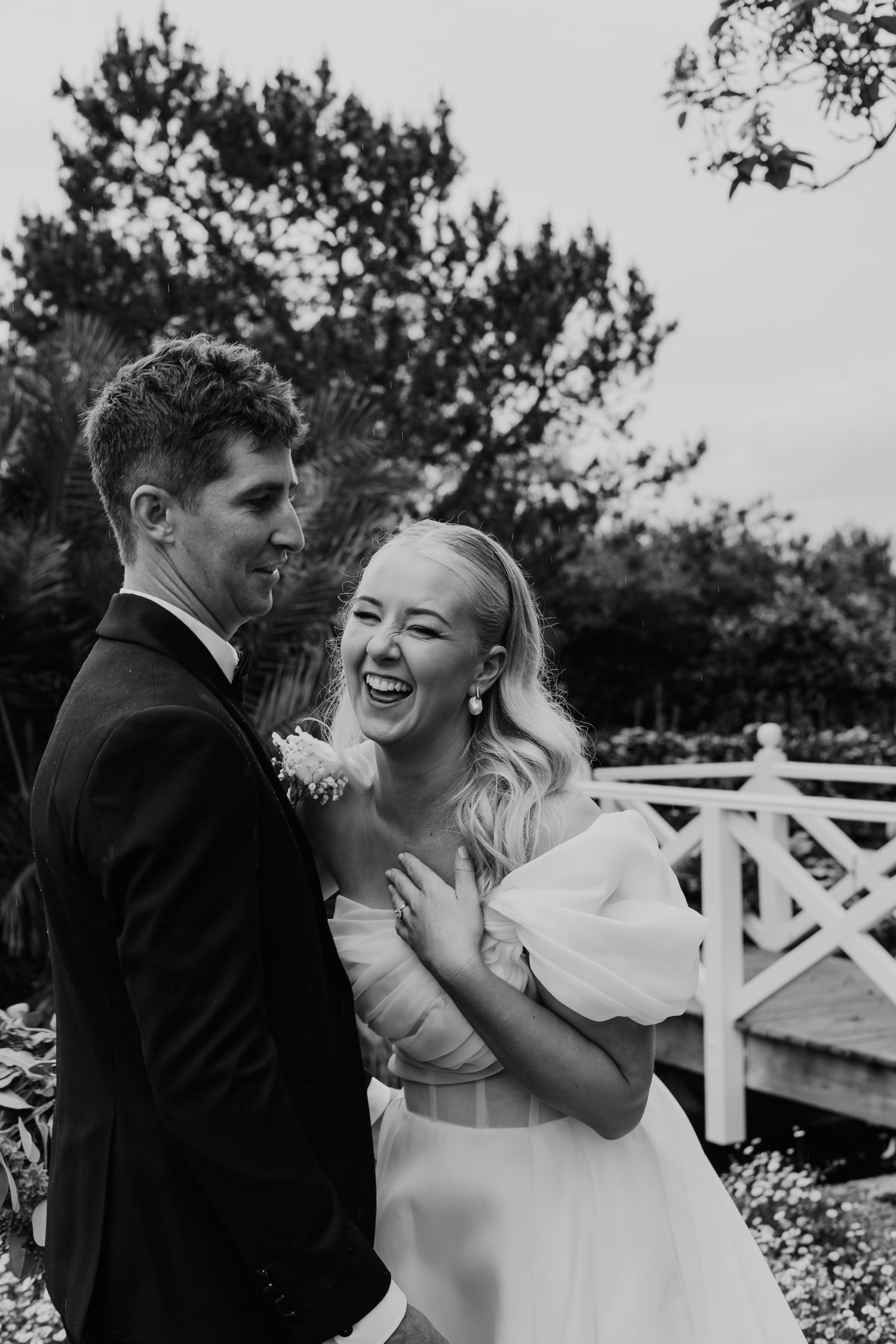 Black and white photo of laughing bride in off-shoulder gown with groom in tuxedo outdoors.