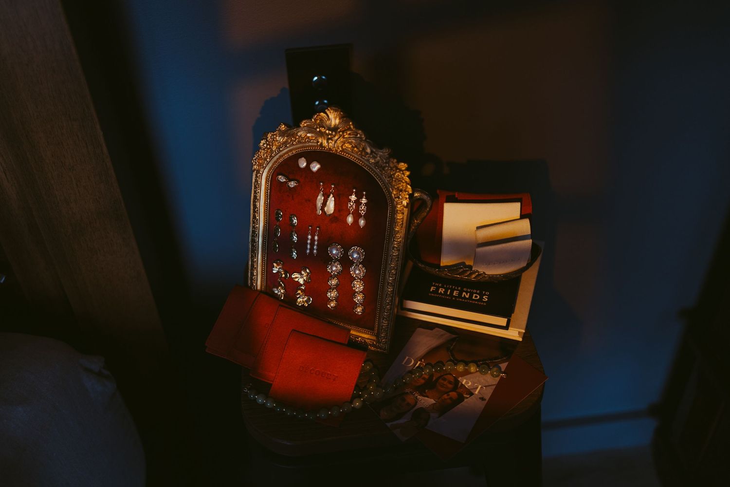 An ornate vintage jewelry box filled with earrings beside a Friends book in moody blue lighting.