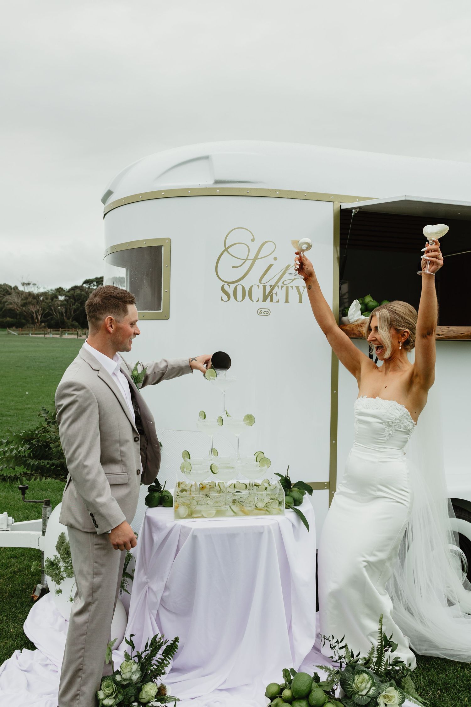 Joyful bride and groom celebrating with champagne tower outside Sip Society mobile bar at wedding.