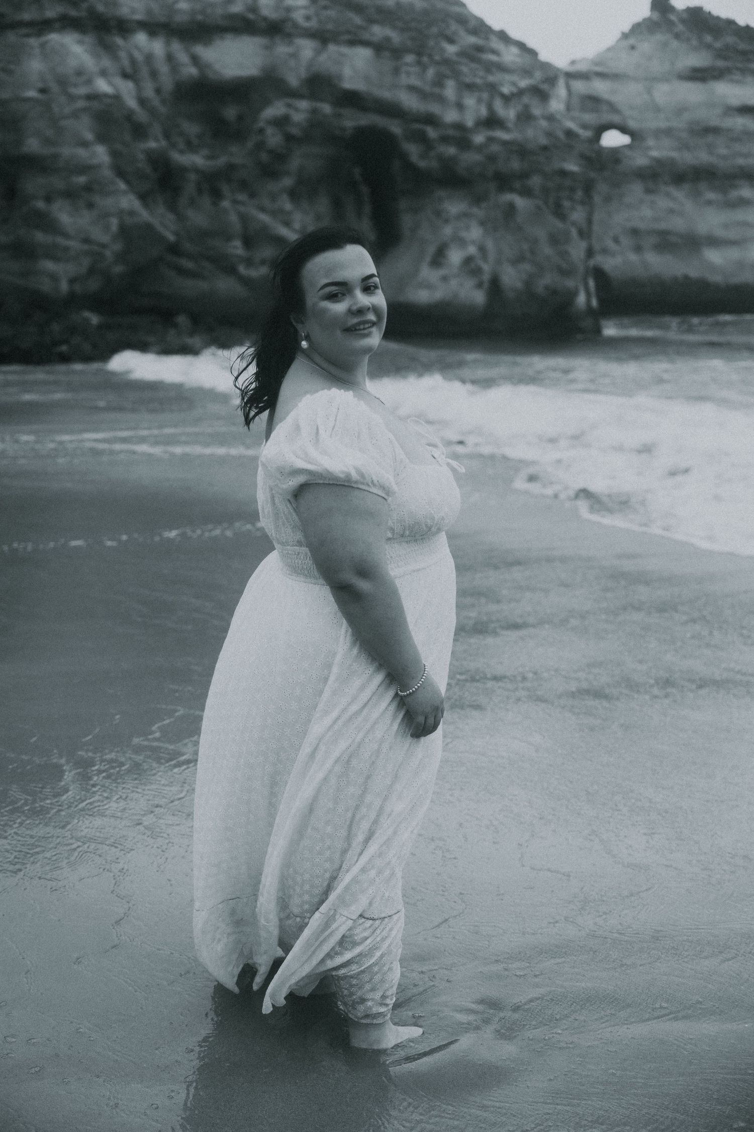 Black and white photo of a woman in a flowing white dress standing in ocean waves near rocky cliffs.