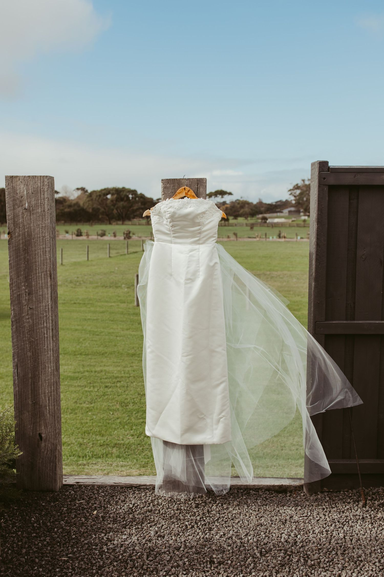Elegant strapless white wedding dress with veil hanging on rustic wooden gate in countryside setting.