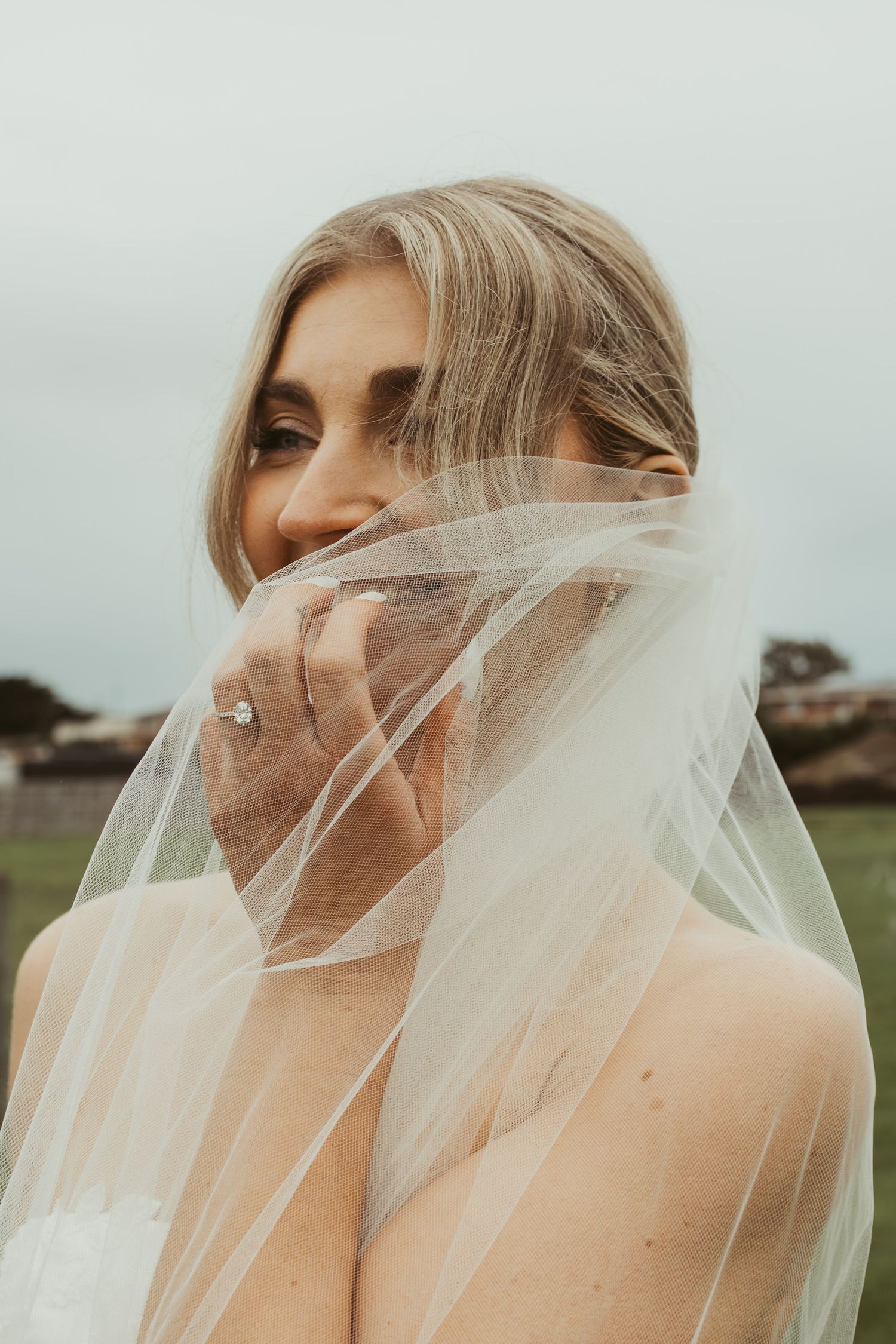 Beautiful blonde bride holding sheer wedding veil across her face, showcasing diamond engagement ring outdoors.