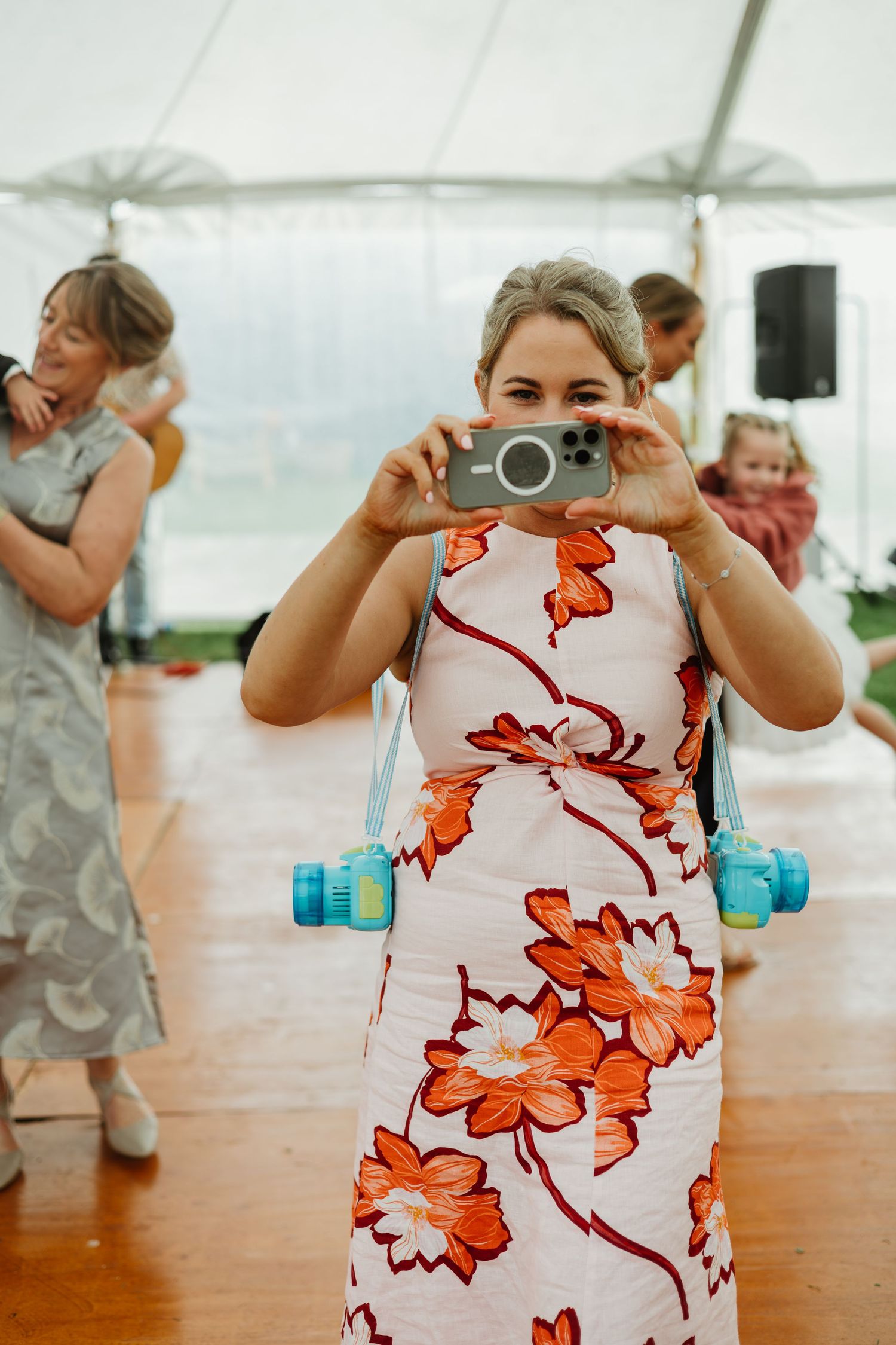 Wedding guest in floral dress photographing with smartphone on wooden dance floor inside white tent.