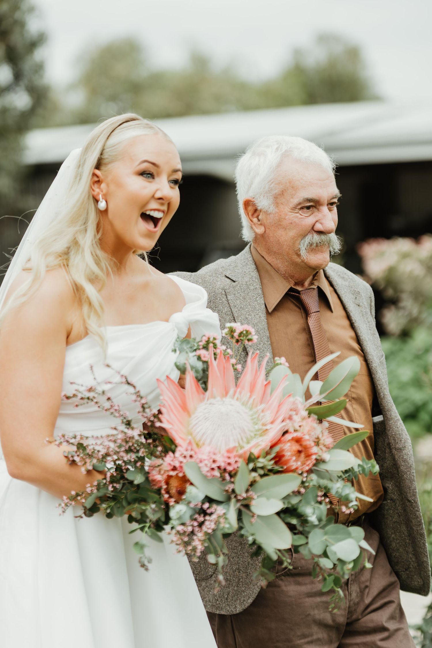 Bride in white gown laughing with elderly man in brown suit, holding tropical bouquet with pink protea outdoors.