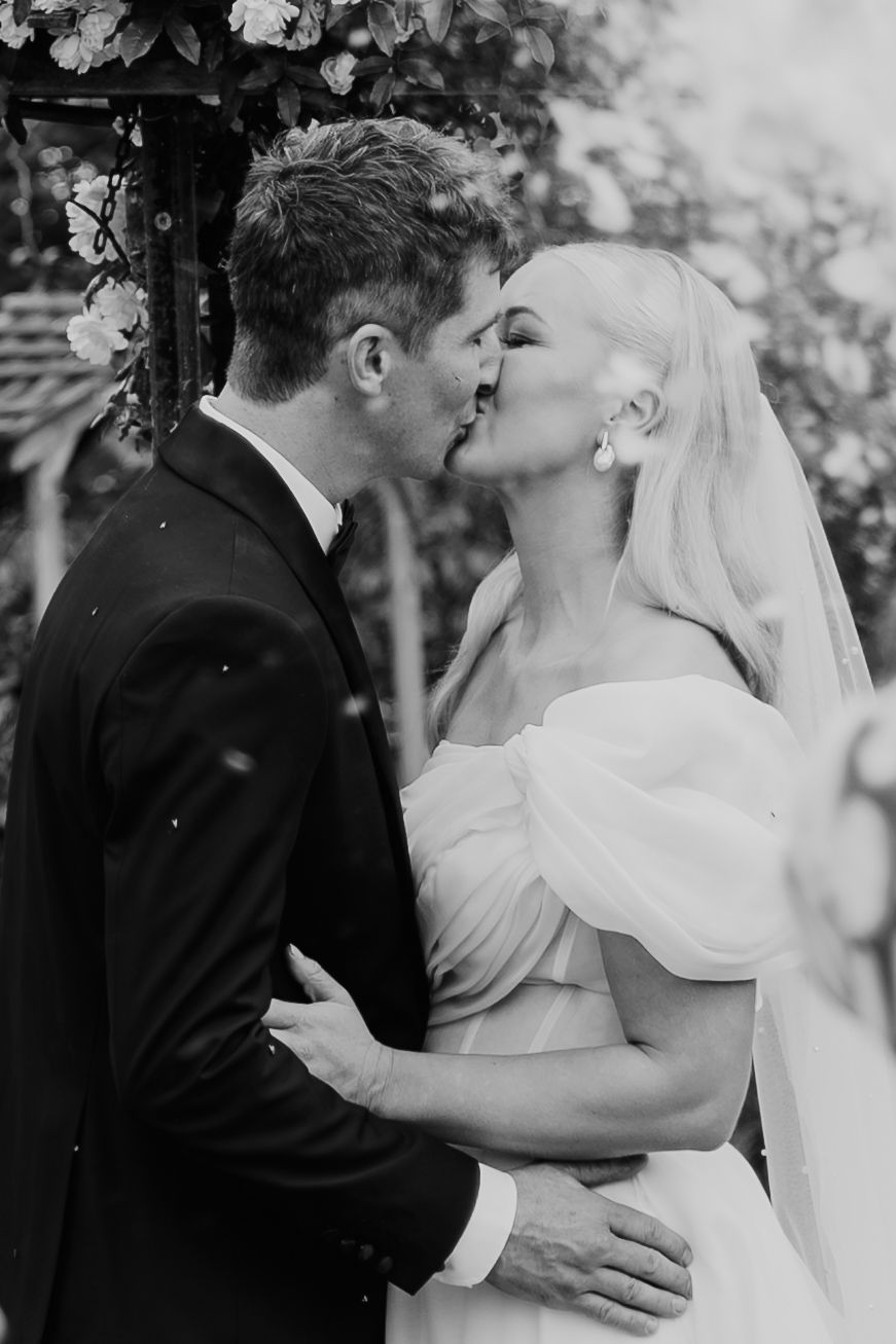 Black and white photo of a bride and groom sharing their first kiss at an outdoor wedding ceremony.