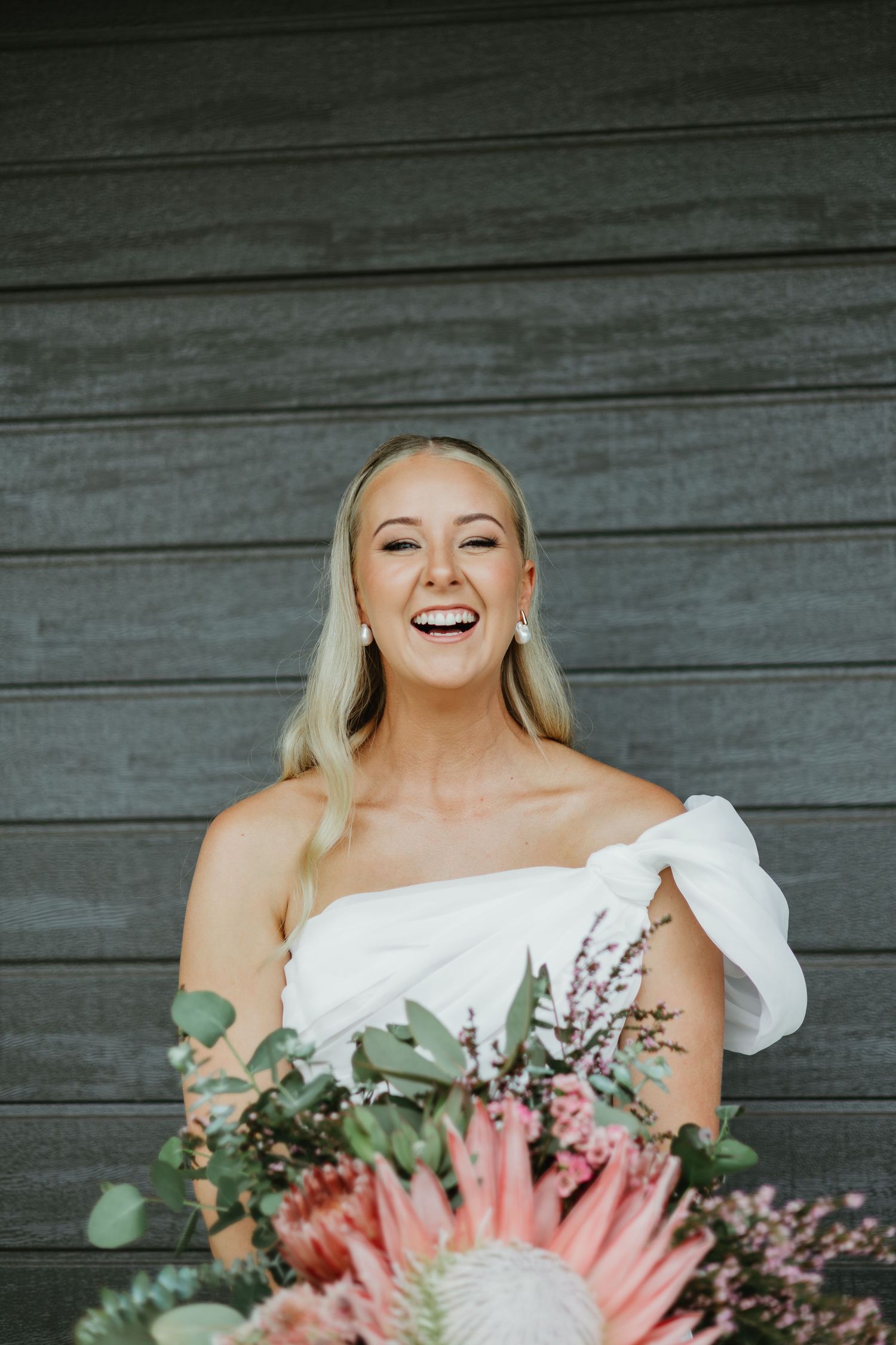Laughing bride in white off-shoulder gown holding a pink protea bouquet with eucalyptus greenery outdoors.