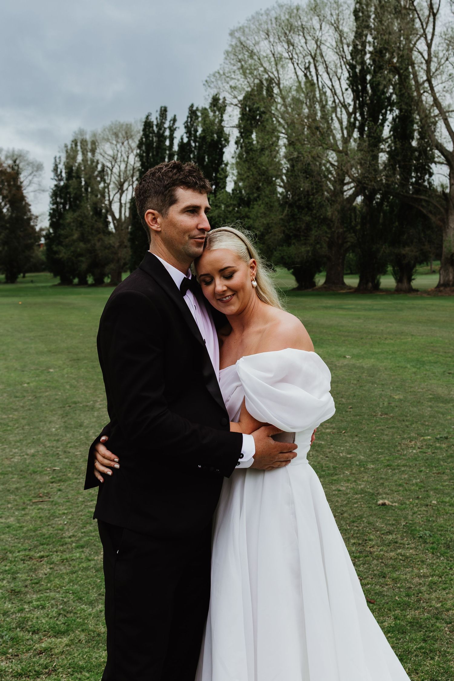 Bride in off-shoulder white gown embracing groom in black tuxedo on green lawn.