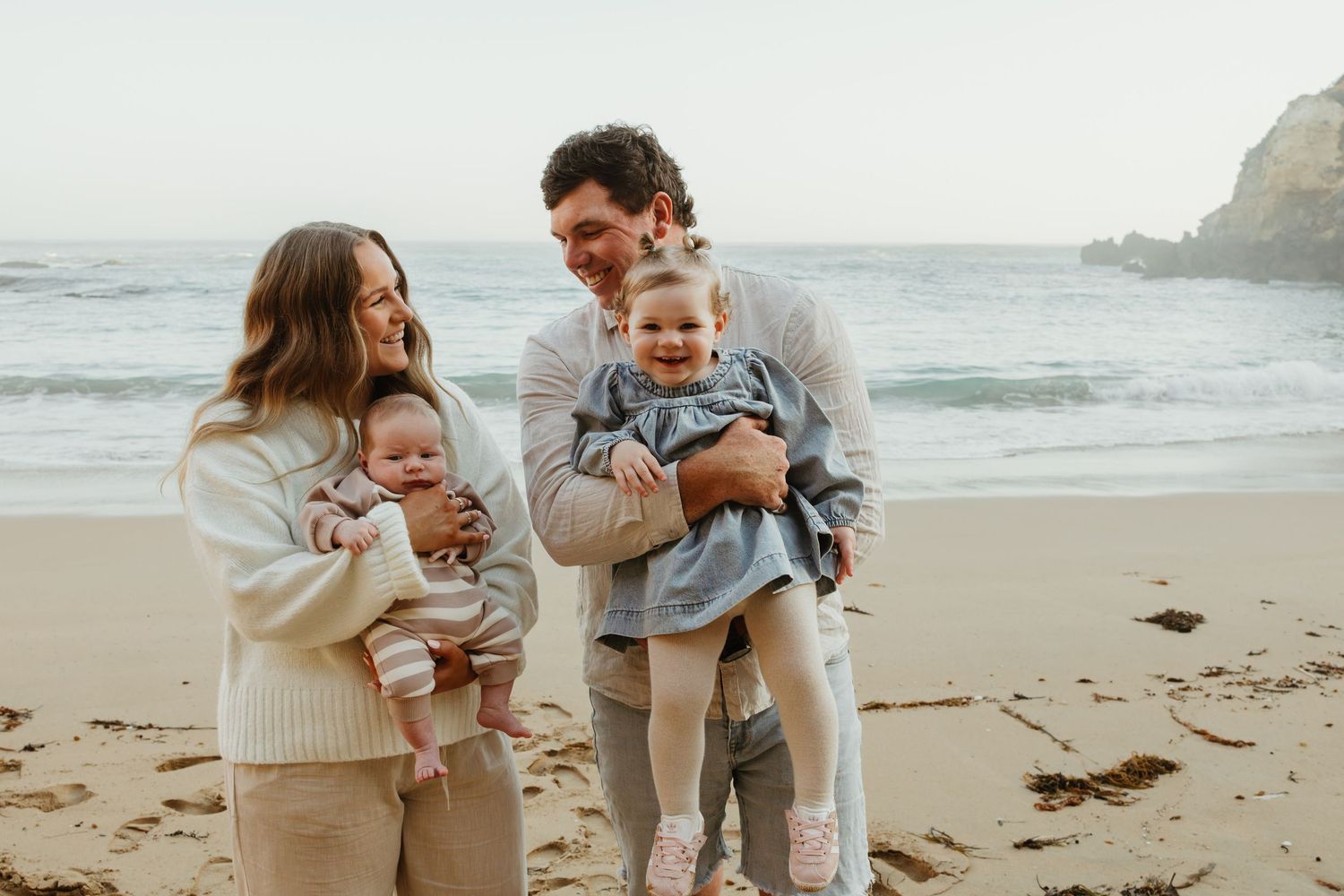 A happy young family with two babies poses on a scenic beach with ocean waves and rocky cliffs in the background.