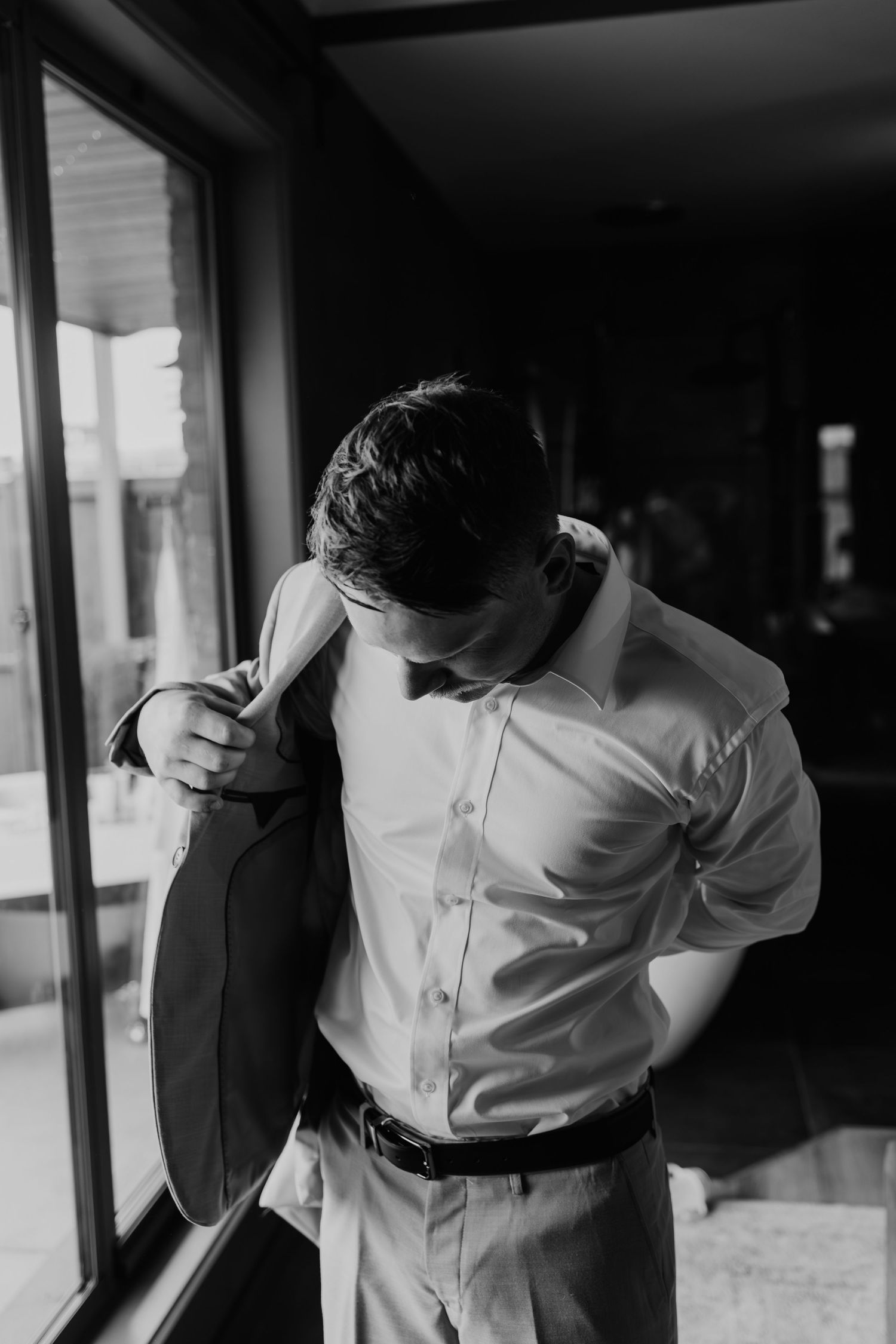 Groom putting on his suit jacket while getting ready near a window on his wedding day.