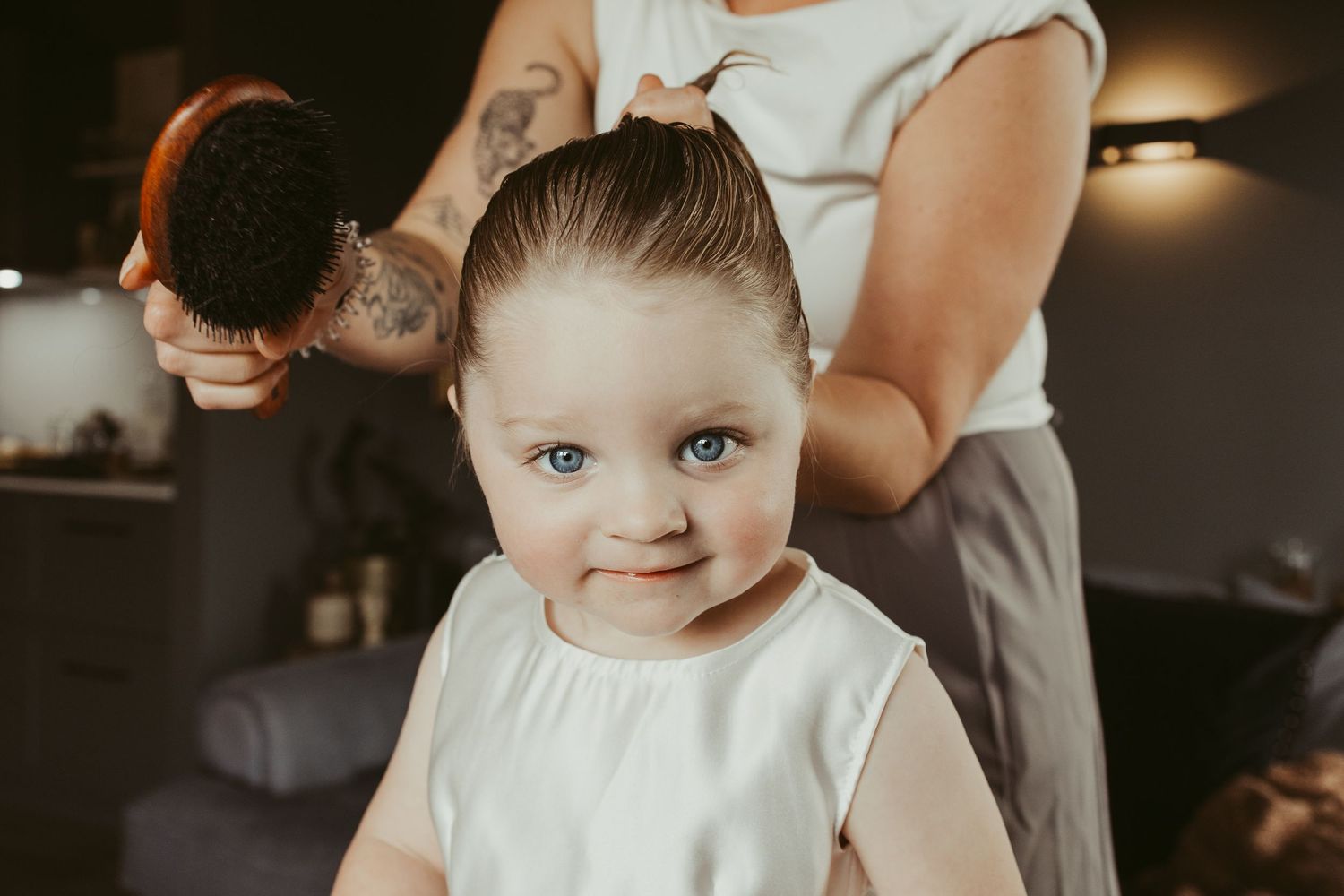 Adorable blue-eyed toddler girl in white dress getting hair styled with round brush.