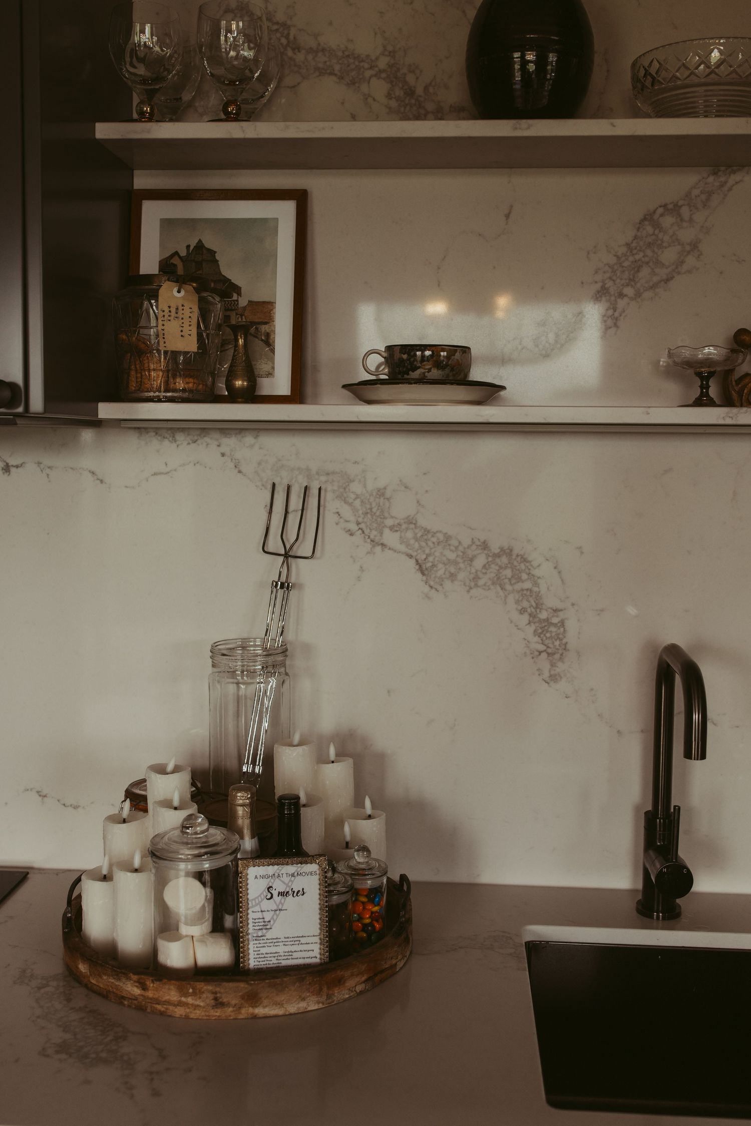 Elegant kitchen counter with marble backsplash, candles on wooden tray, decorative shelves with vases and artwork.