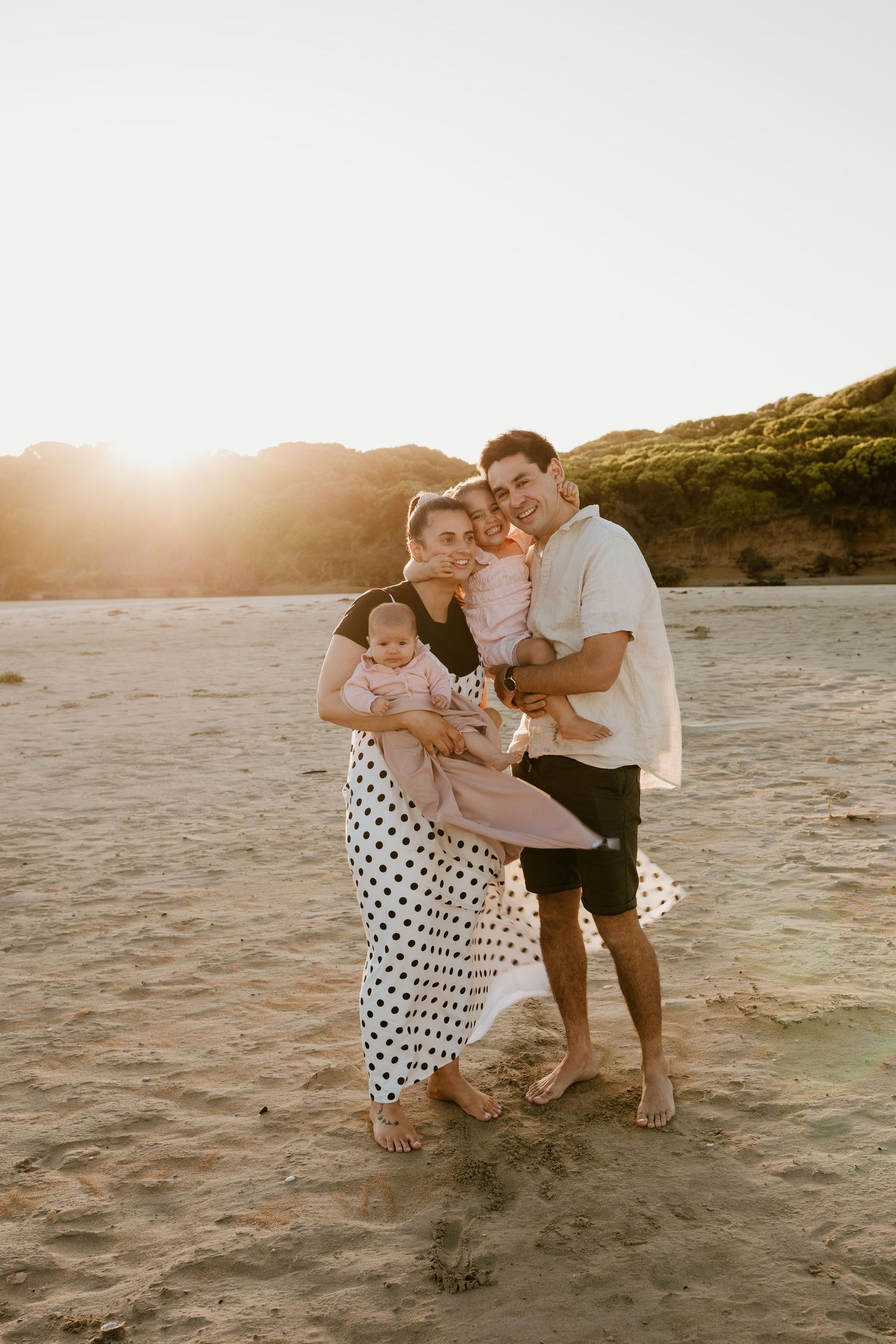 A happy family of four poses on a sandy beach at golden sunset, parents holding two young children.