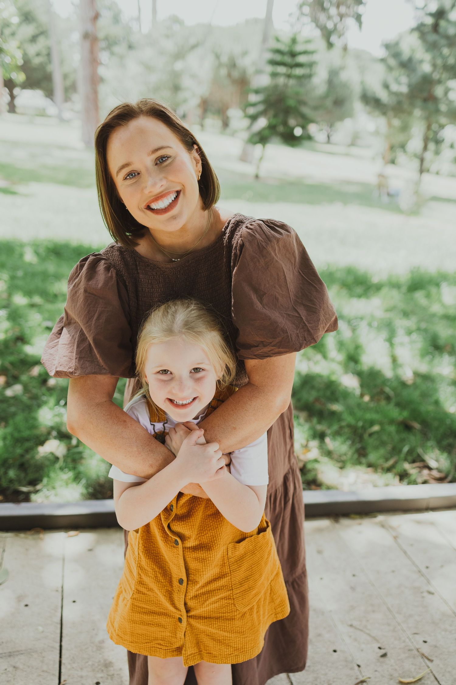 Smiling brunette mother hugs her young blonde daughter in a yellow skirt outdoors in a park.