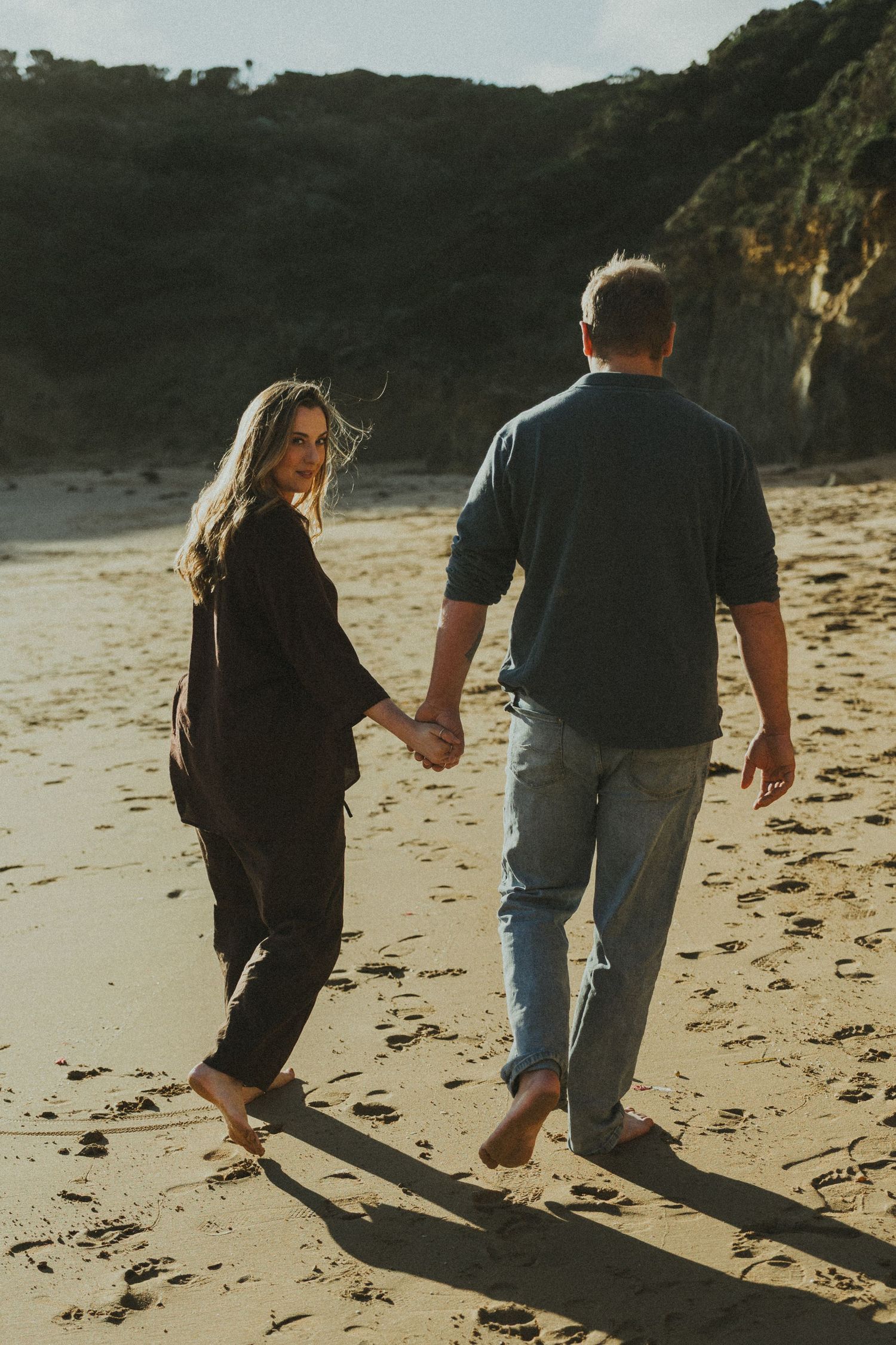 A couple holds hands while walking barefoot on a sandy beach with rocky cliffs at sunset.