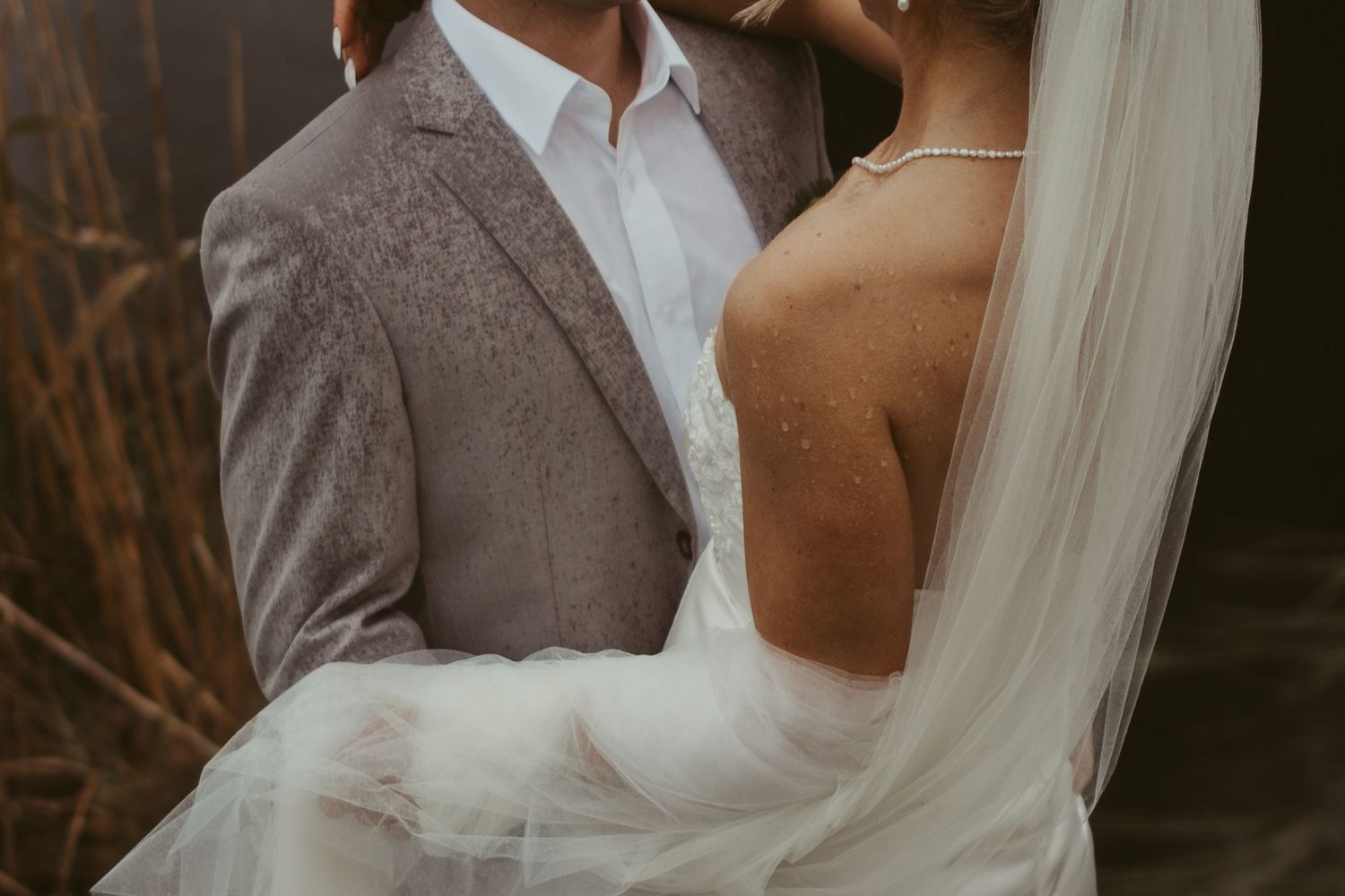 Romantic wedding couple embracing outdoors, bride in white gown with veil, groom in gray suit.