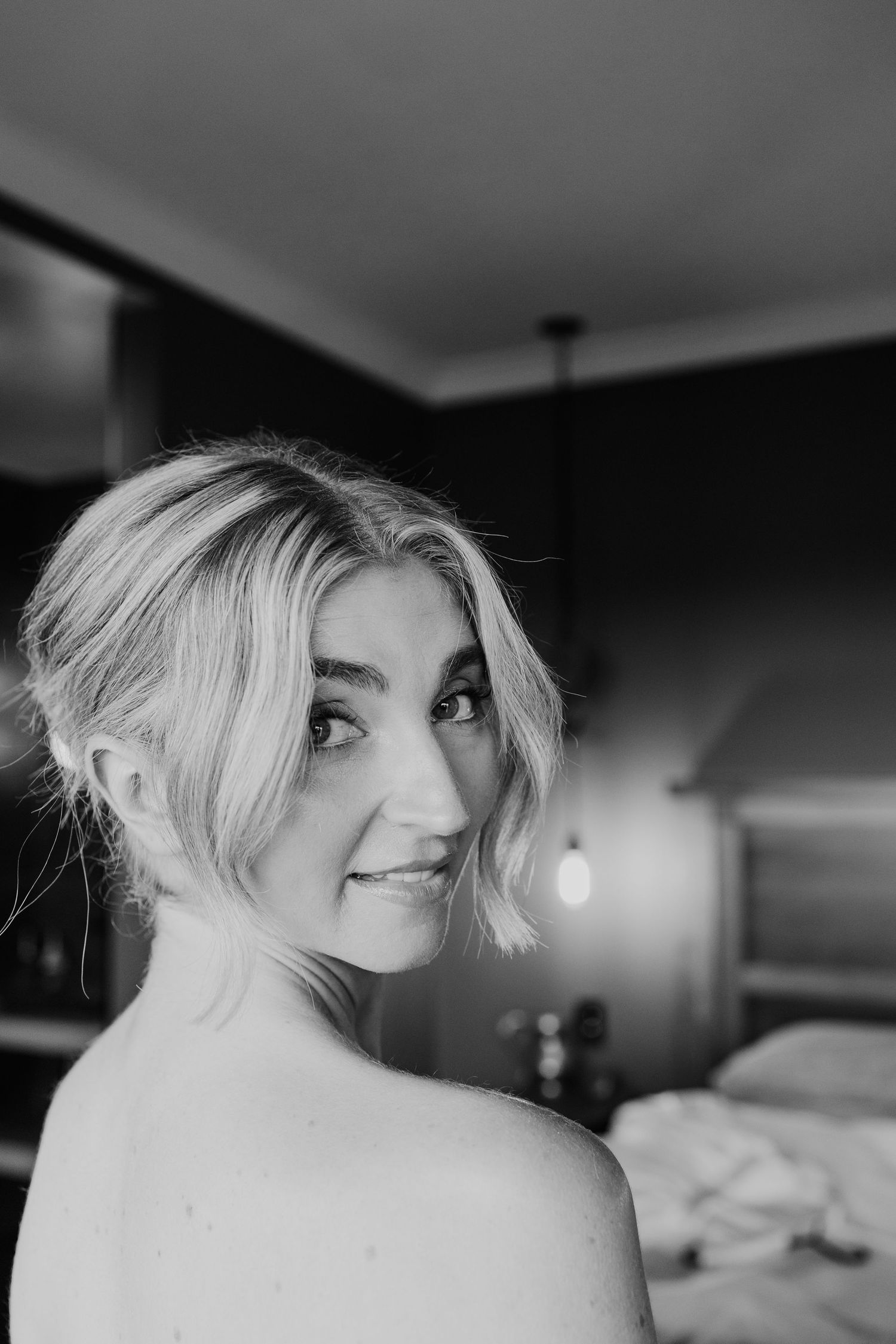 Black and white portrait of smiling blonde bride with short hair looking over bare shoulder.