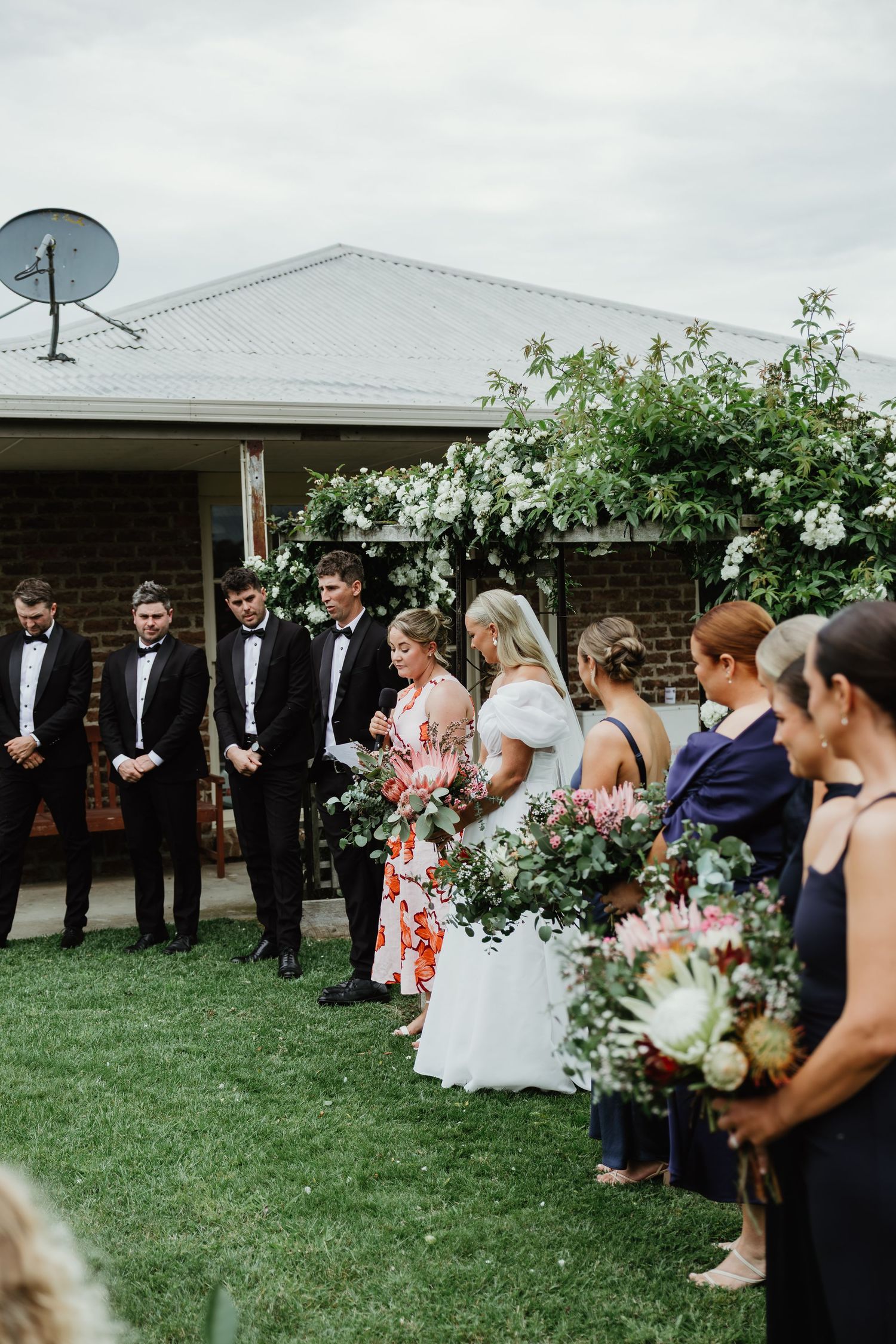 Wedding party standing outdoors by a floral arch at a country farm ceremony.