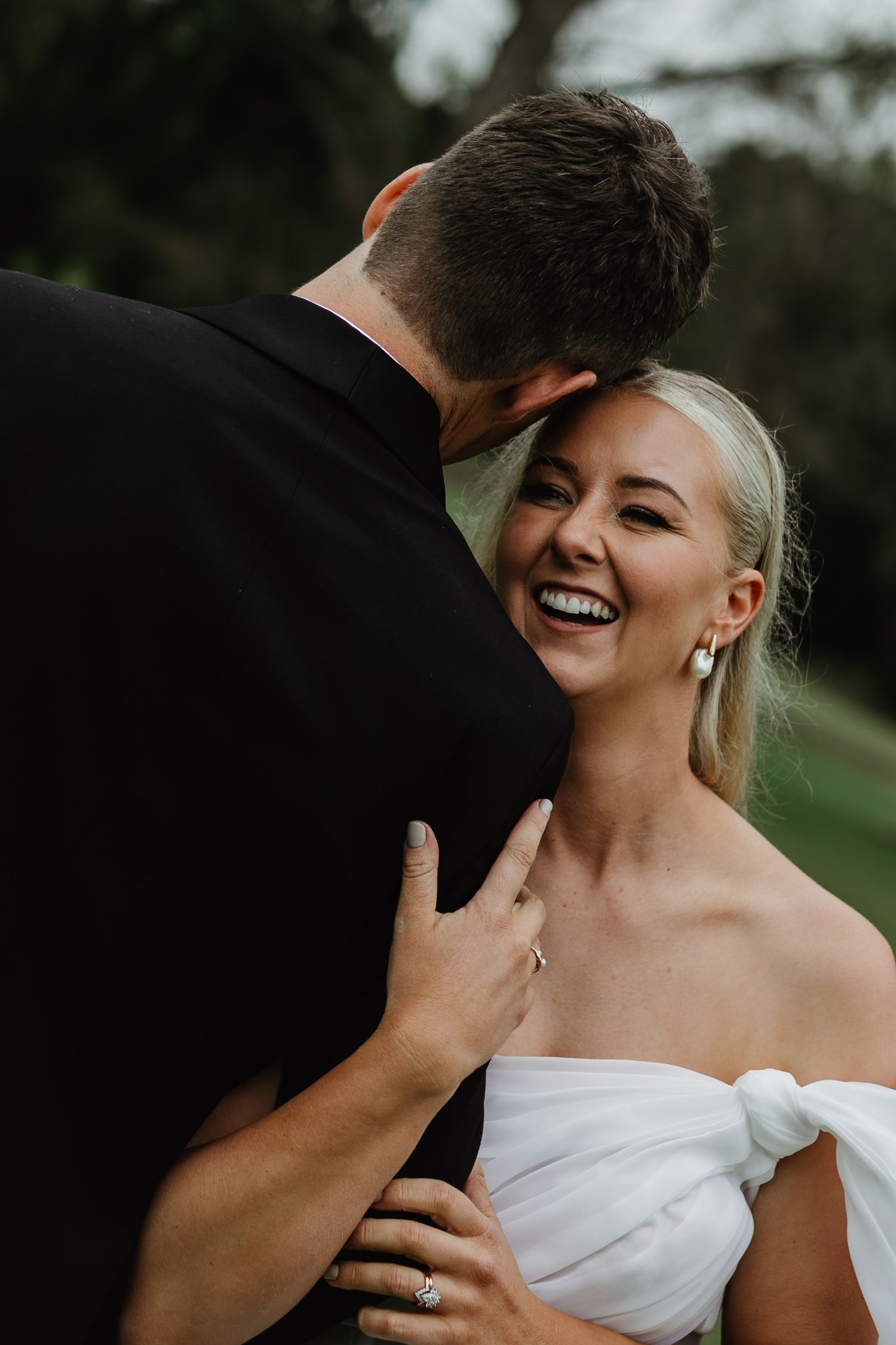 Laughing bride in off-shoulder gown embraces groom showing diamond engagement ring.