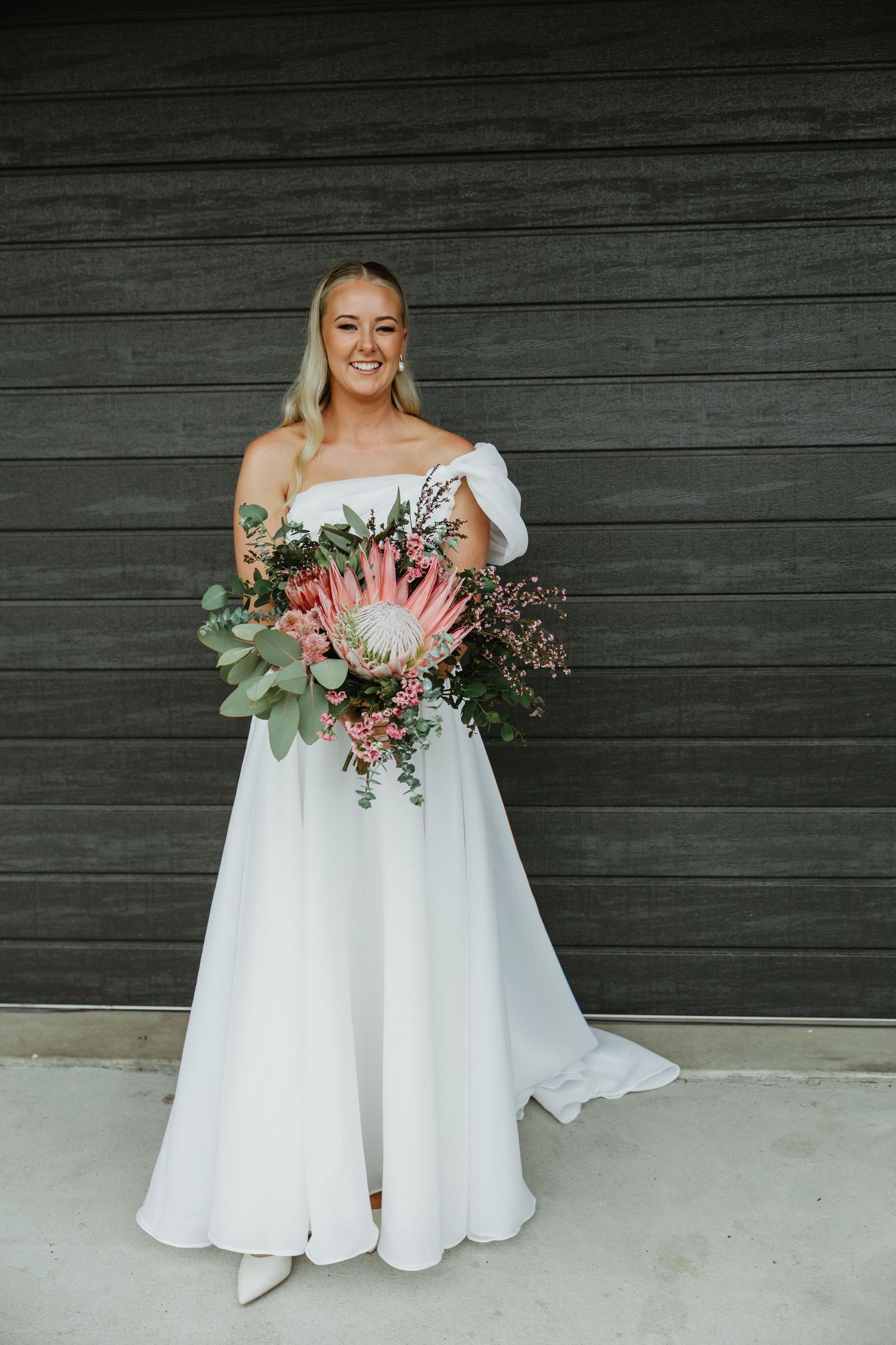 Smiling bride in off-shoulder white gown holding a large native protea bouquet with eucalyptus foliage.