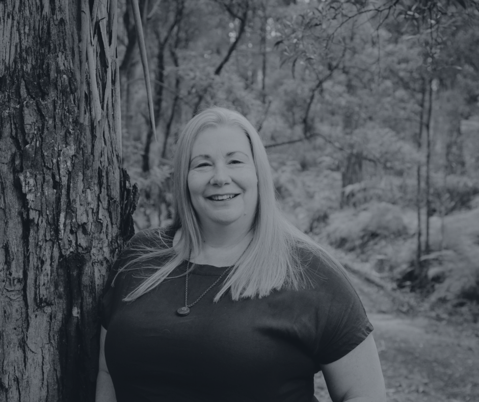 Woman with blonde hair smiling outdoors, leaning against a tree in a lush forest setting, wearing a dark top.
