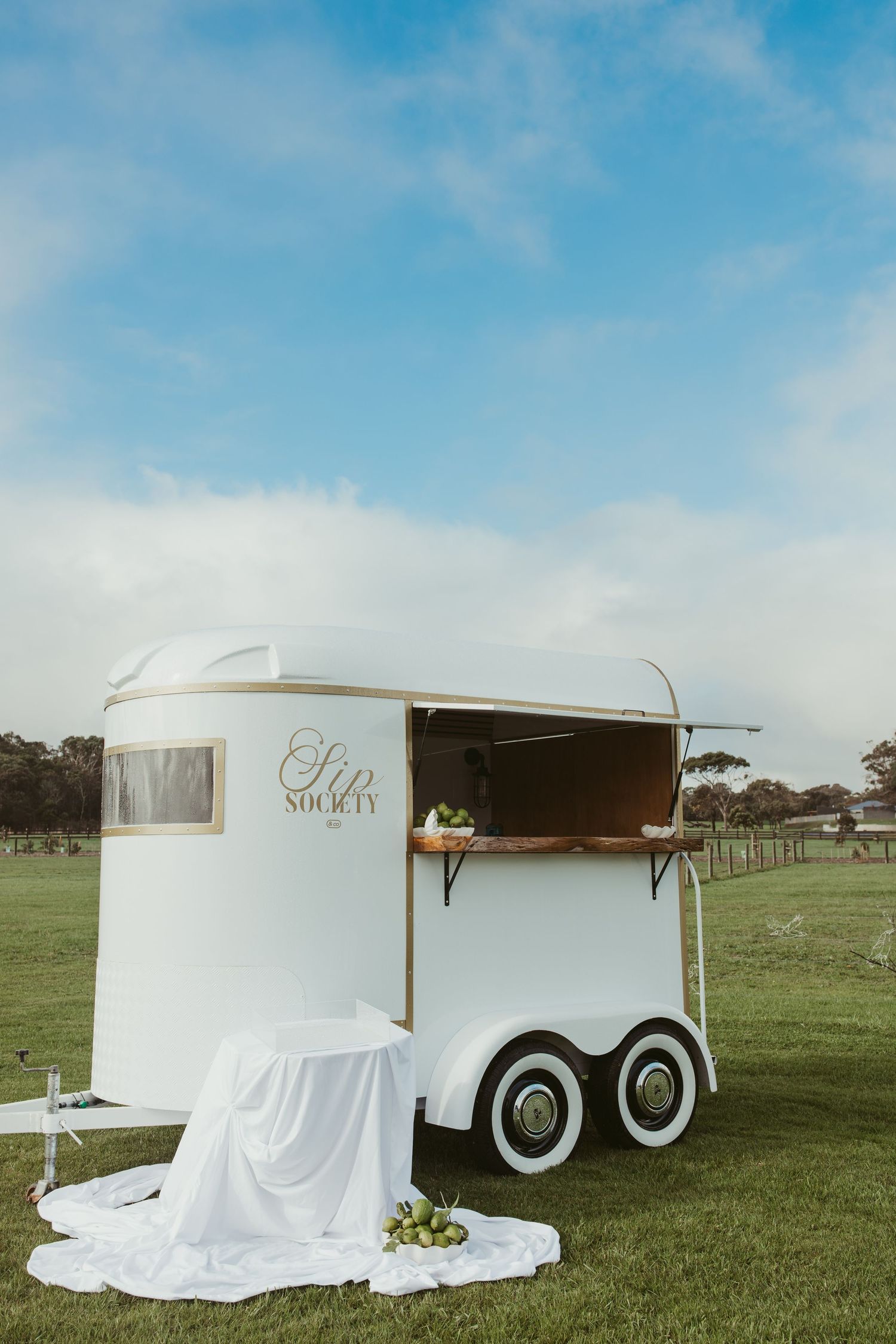 White horse trailer bar named Sip Society with gold accents on green field under blue sky.
