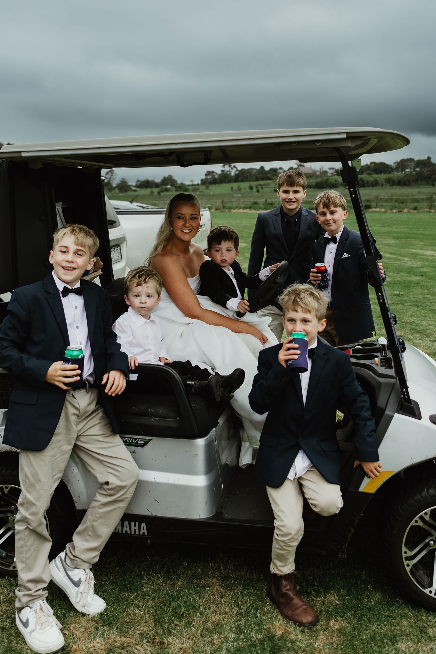 Bride in white gown poses with young boys in suits on golf cart at outdoor wedding venue.