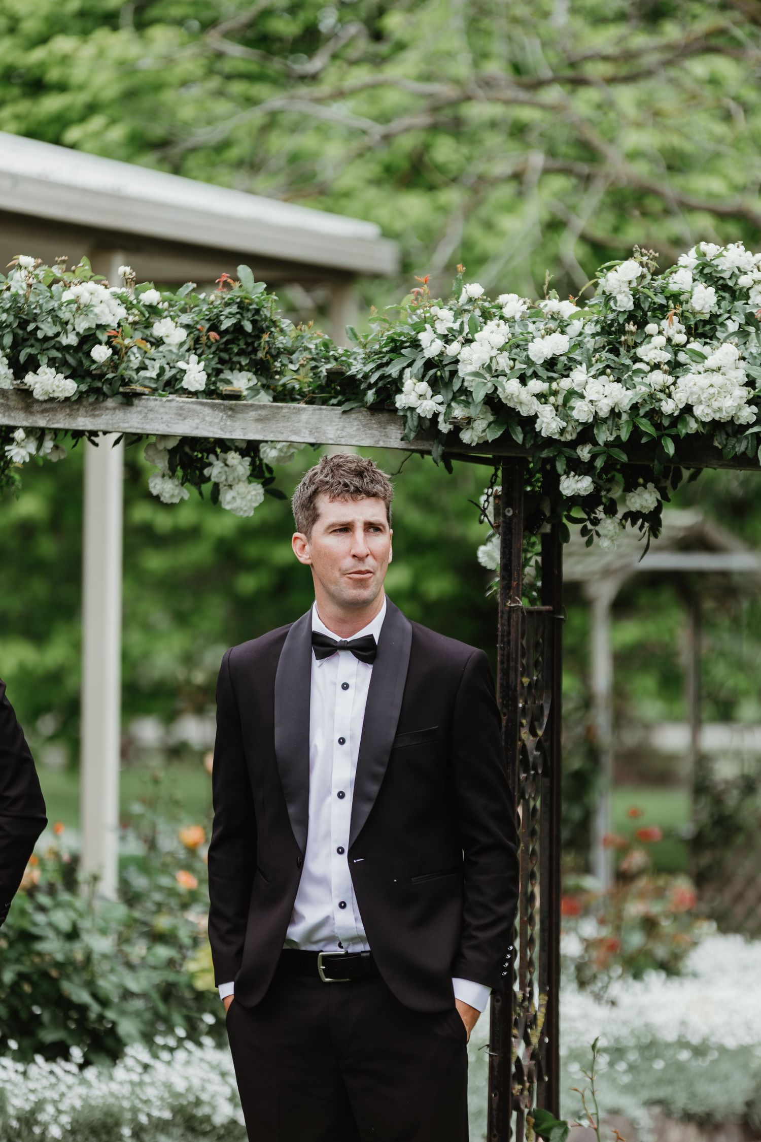 Groom in black tuxedo standing under floral arch with white roses at outdoor wedding ceremony.