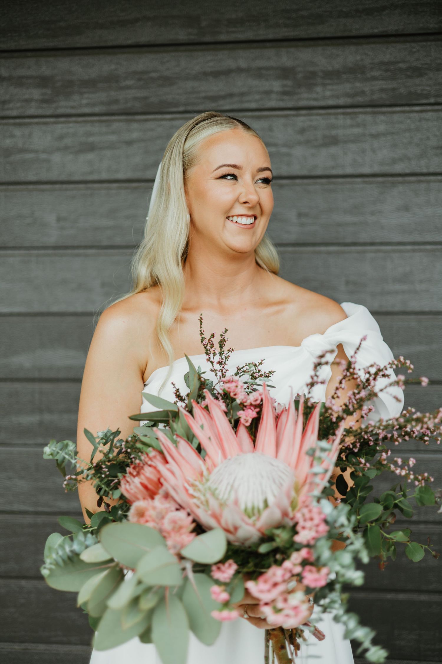 Smiling blonde bride holds large pink protea bouquet in off-shoulder white wedding dress.