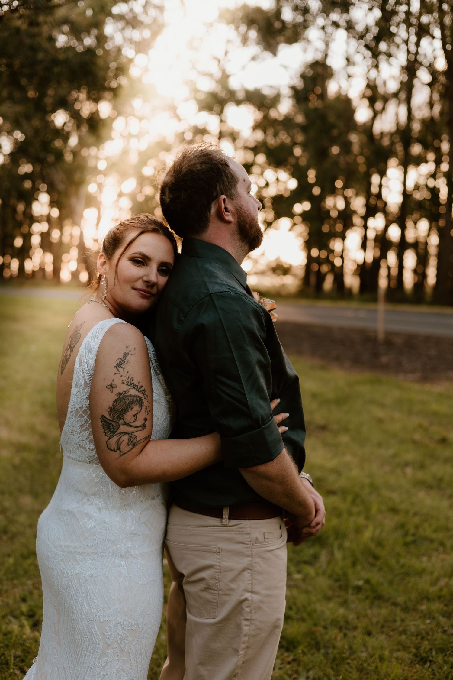 Bride in white lace dress with tattoo leans on groom in green shirt at golden hour outdoor wedding portrait.