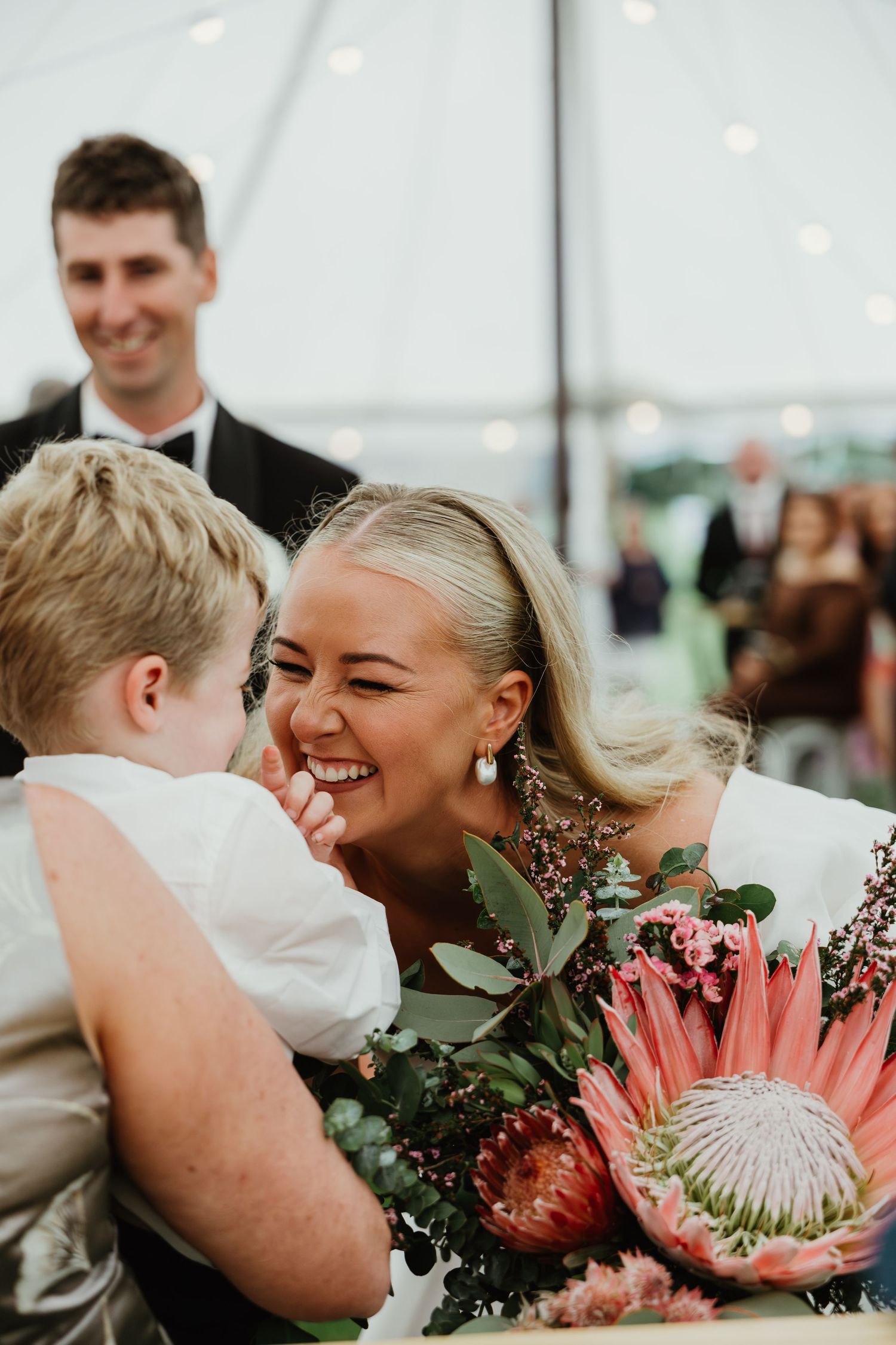Smiling bride leans in to interact with young child while holding large pink protea bouquet at reception.