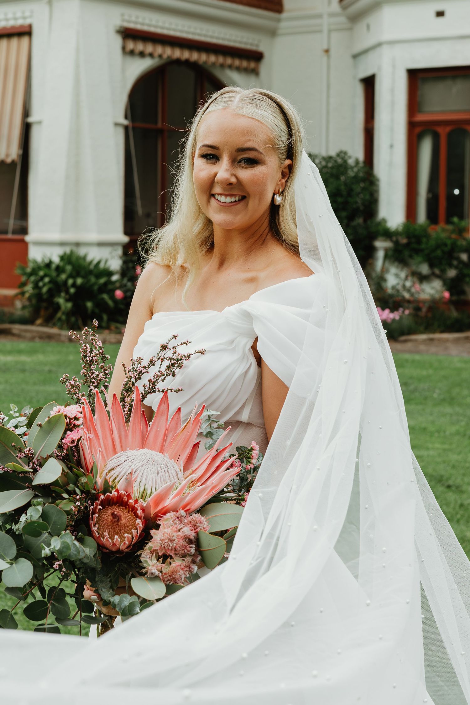 Smiling blonde bride in white gown holding large pink protea bouquet with veil flowing.