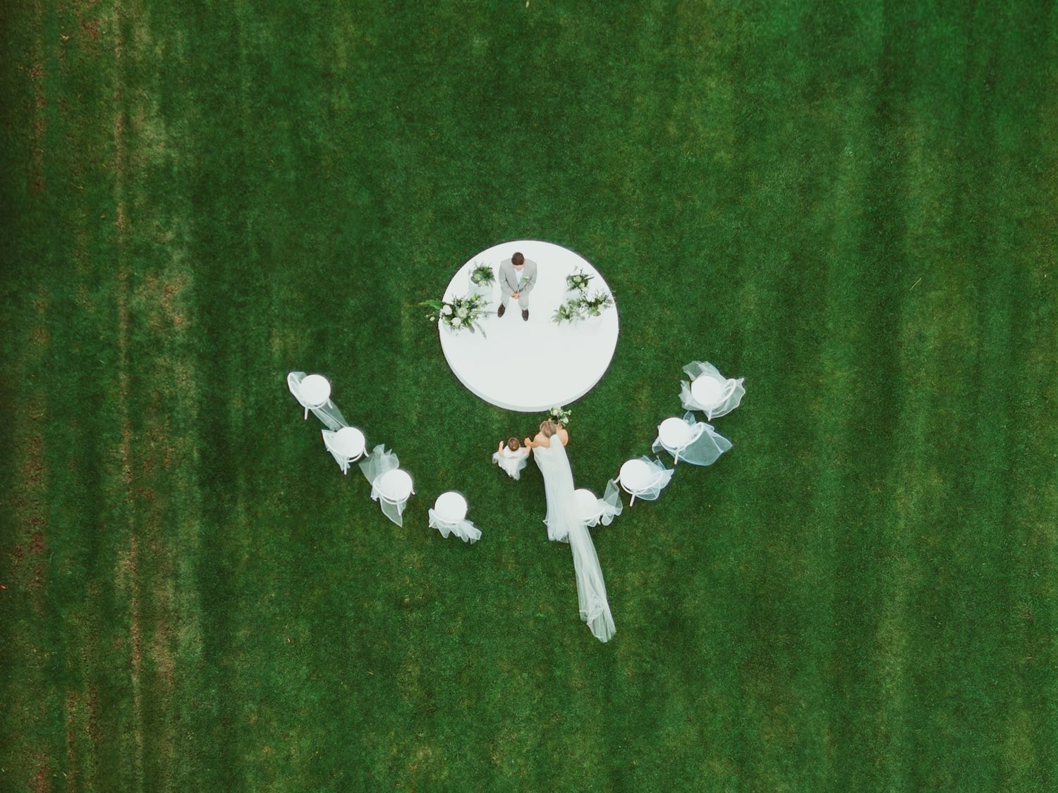 Aerial drone view of intimate outdoor wedding ceremony on circular white platform surrounded by green grass.