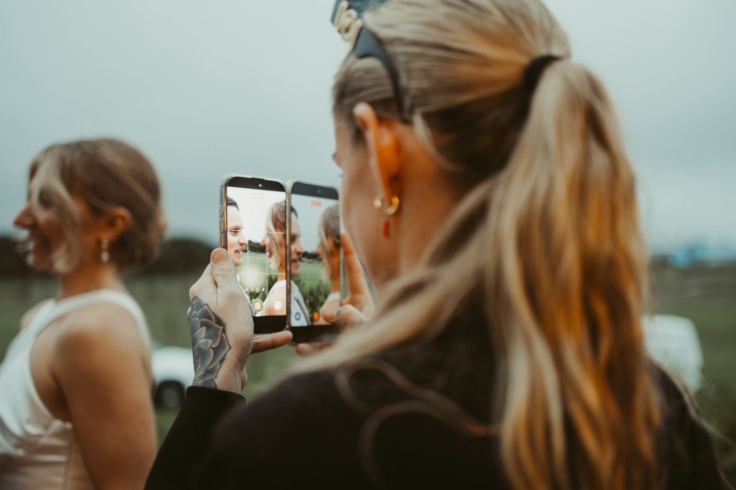 A blonde woman photographs a bride with her smartphone at an outdoor wedding celebration.