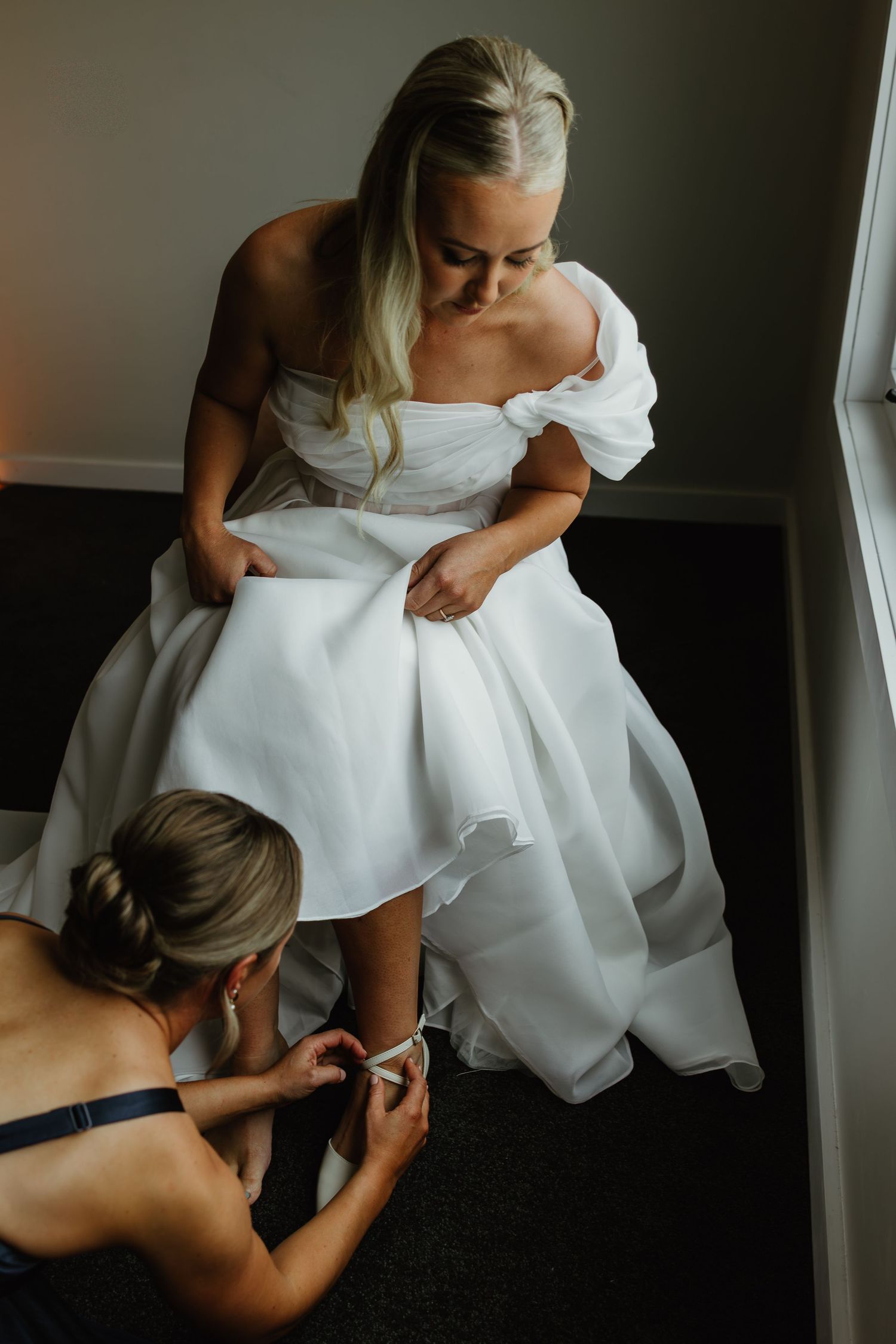 Bridesmaid helping bride with shoe while wearing elegant white wedding dress.